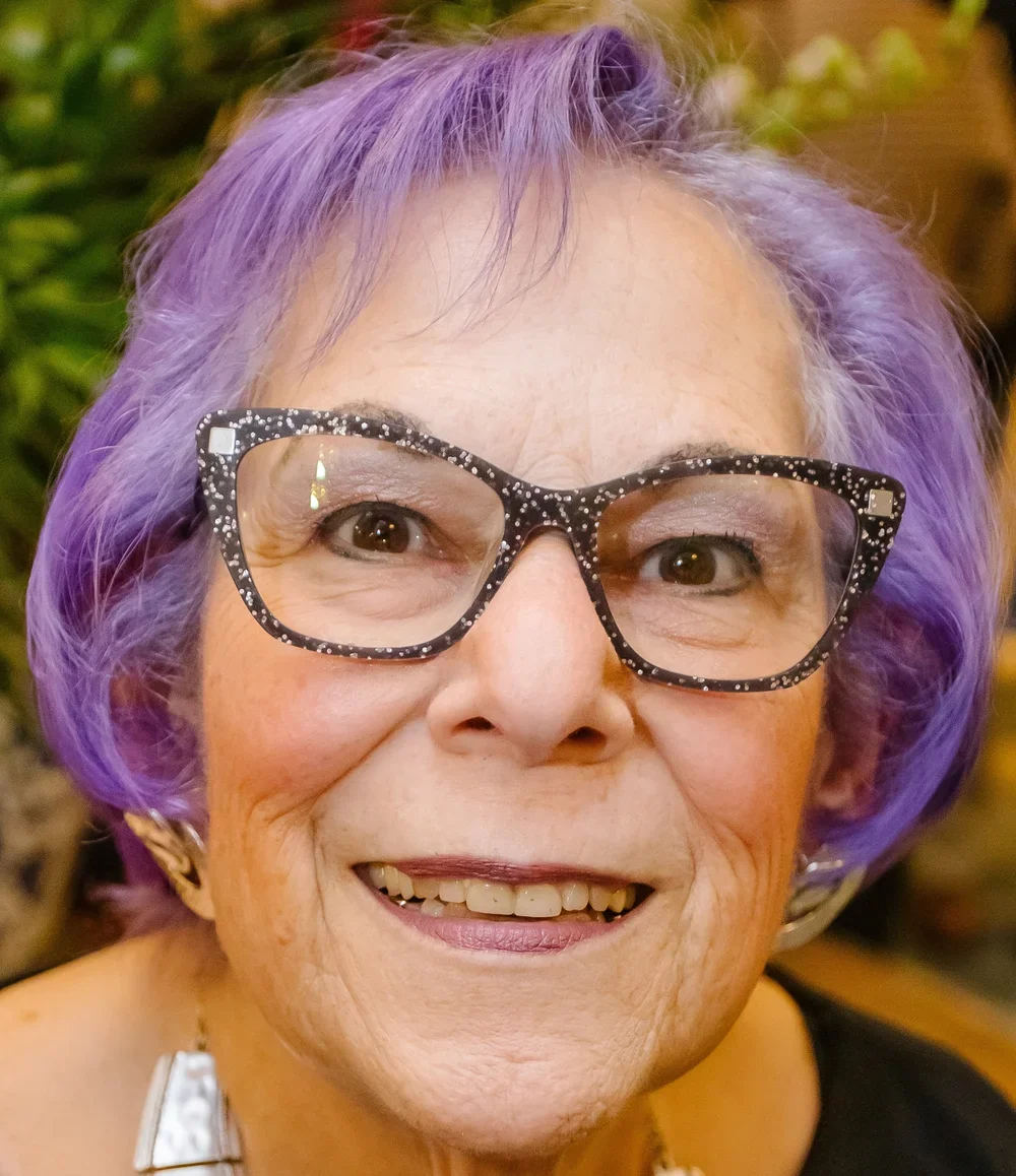 Close-up of a smiling elderly woman with short purple hair, wearing large black glasses with white polka dots, earrings, and a statement necklace.