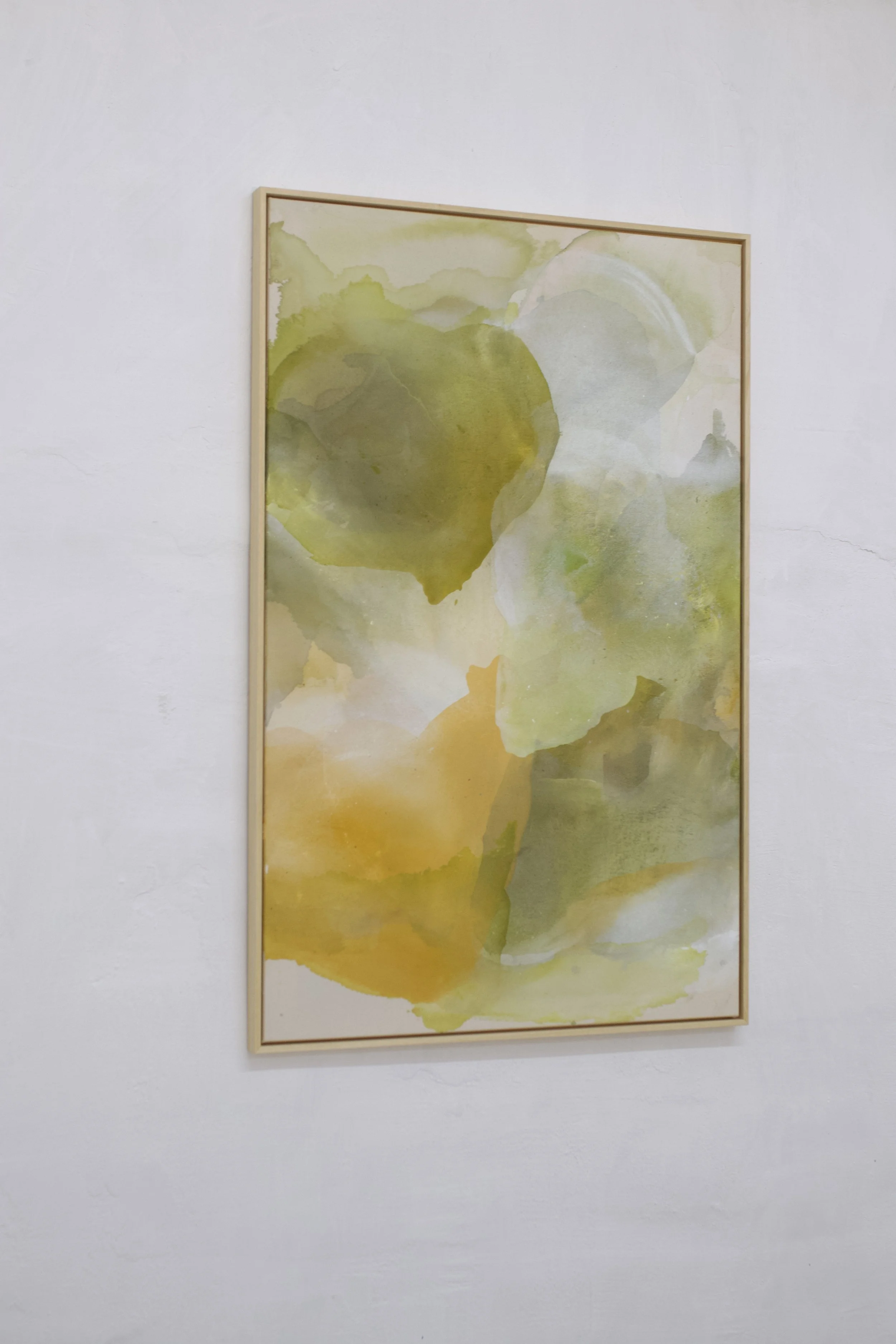 Abstraktes Wasserfarbenbild in Gelb- und Grüntönen in einem Rahmen hängt an einer weißen Wand.