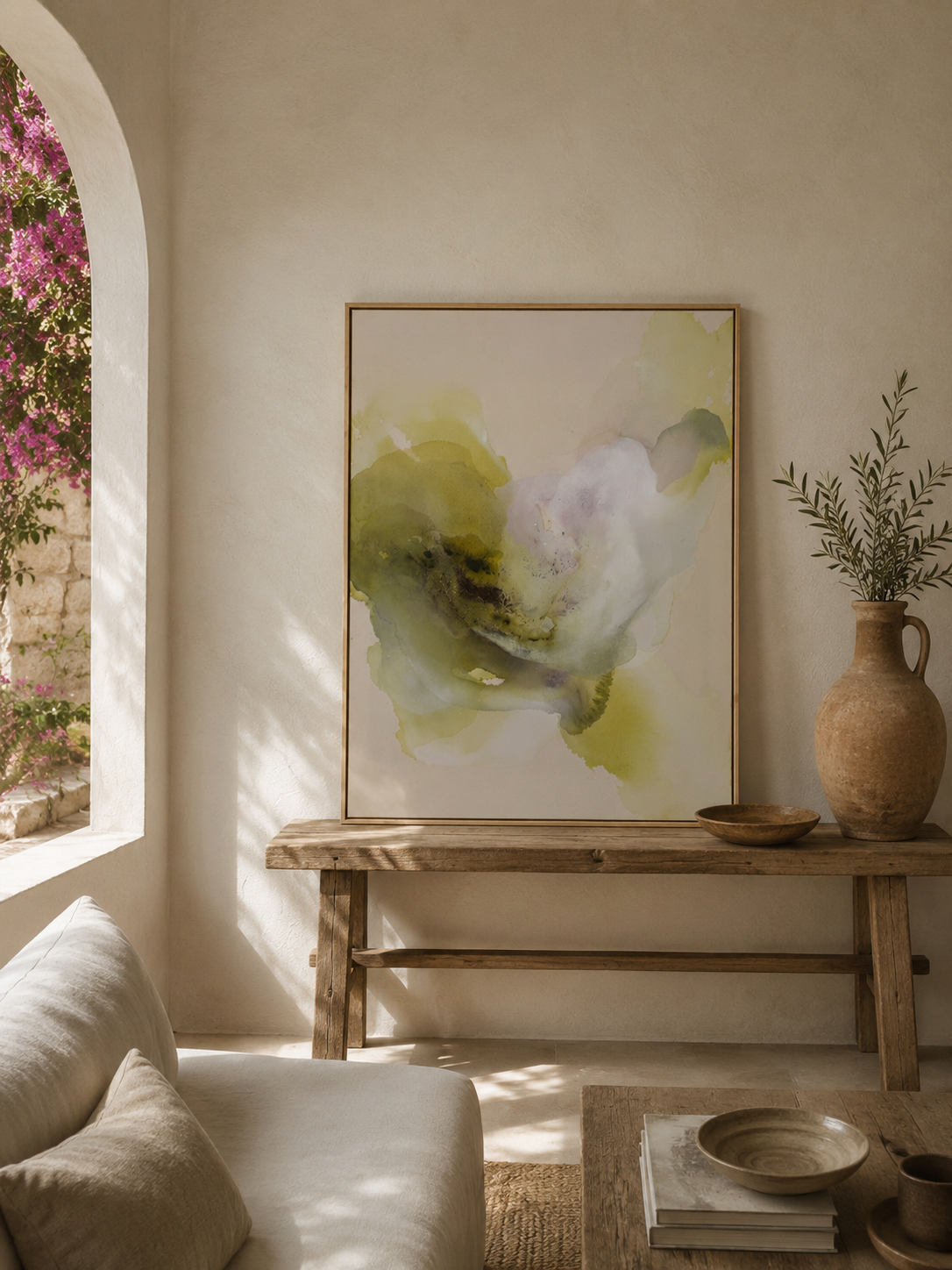 Ein Wohnzimmer mit einem abstrakten Wandbild in Grüntönen, einer Vase mit grünen Zweigen auf einem Holztablett, einem weißen Sofa mit Kissen, einem Fenster mit Blick auf eine blühende Pflanze und einem Couchtisch mit Büchern und Schalen.
