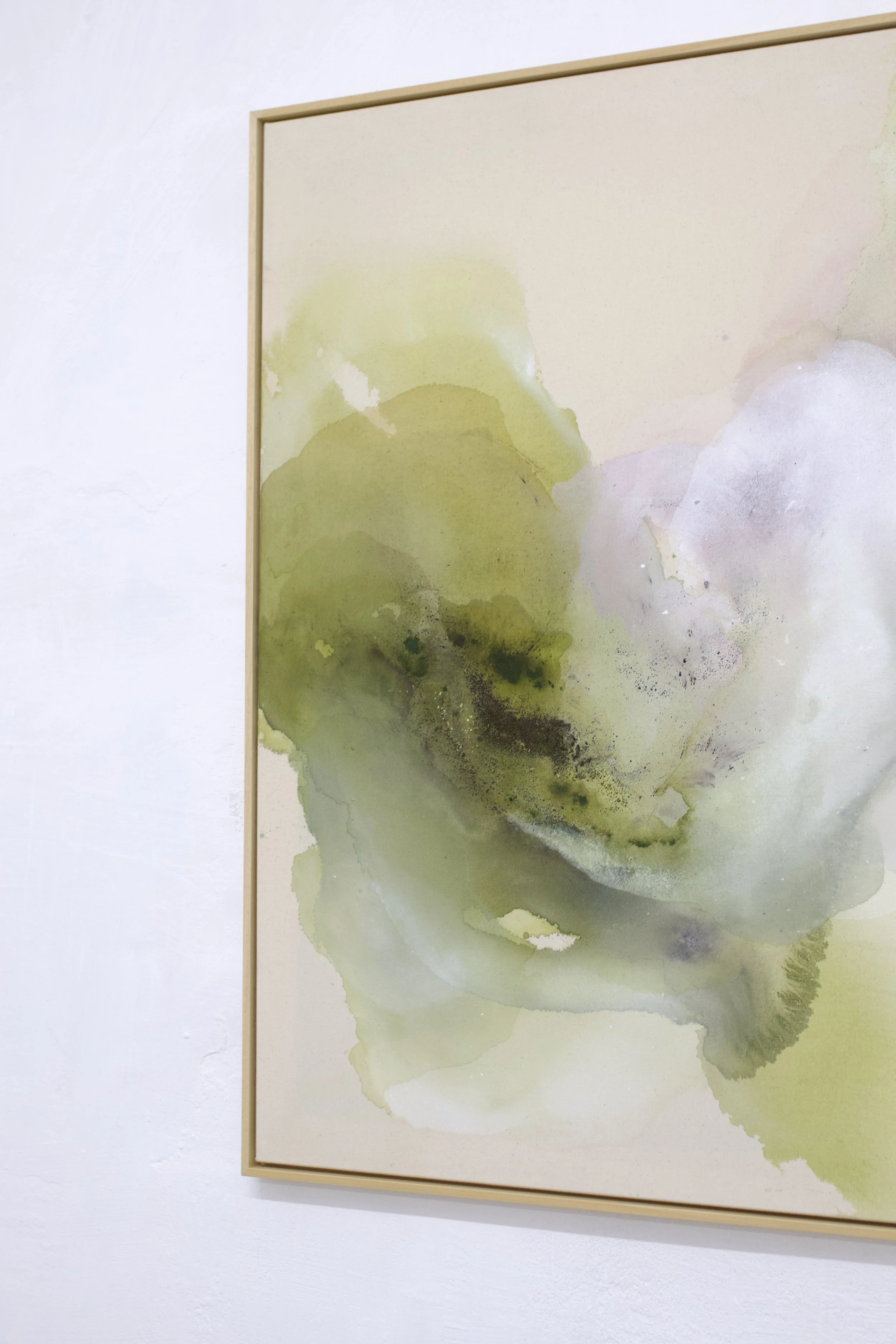 Aquarellgemälde mit grünen und lila Farbverläufen in einem hellen Holzrahmen, an einer weißen Wand.