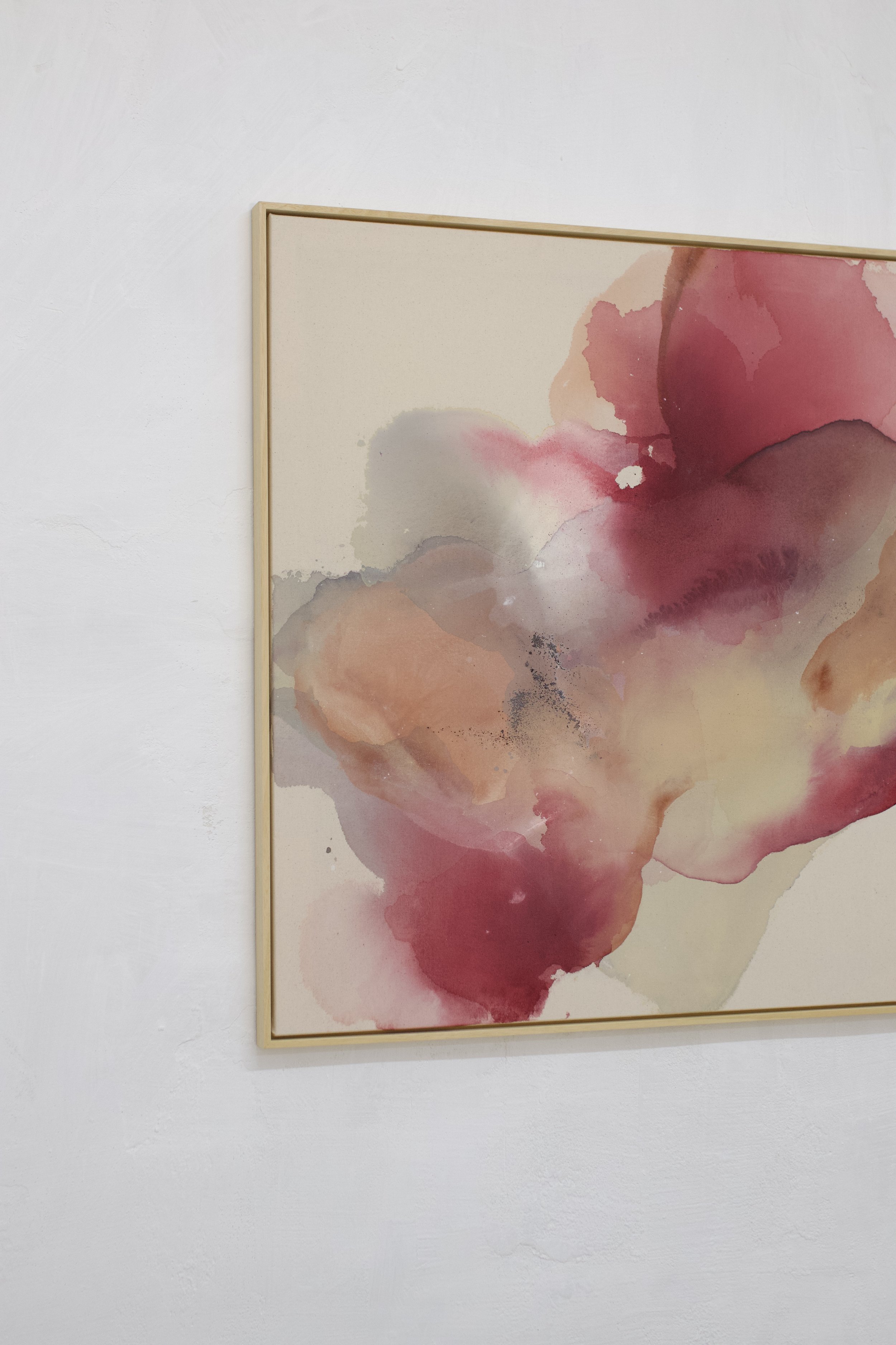 Eine abstrakte Aquarellmalerei mit roten, pinken und beige Tönen, eingerahmt in einen dünnen Holzrahmen, an einer weißen Wand.
