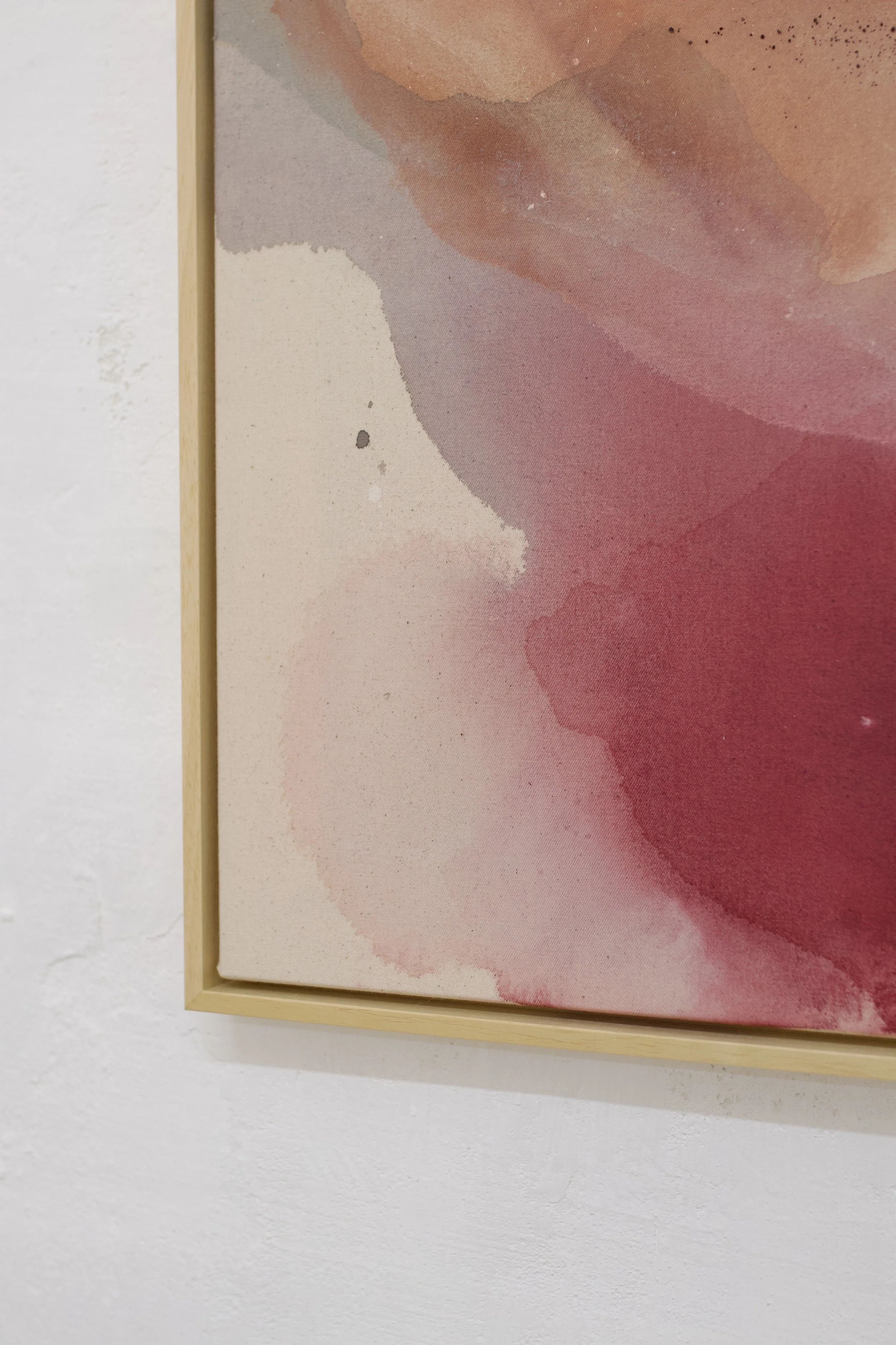 A close-up image of an abstract watercolor painting with shades of pink, peach, and purple, framed in light wood, hanging on a white wall.