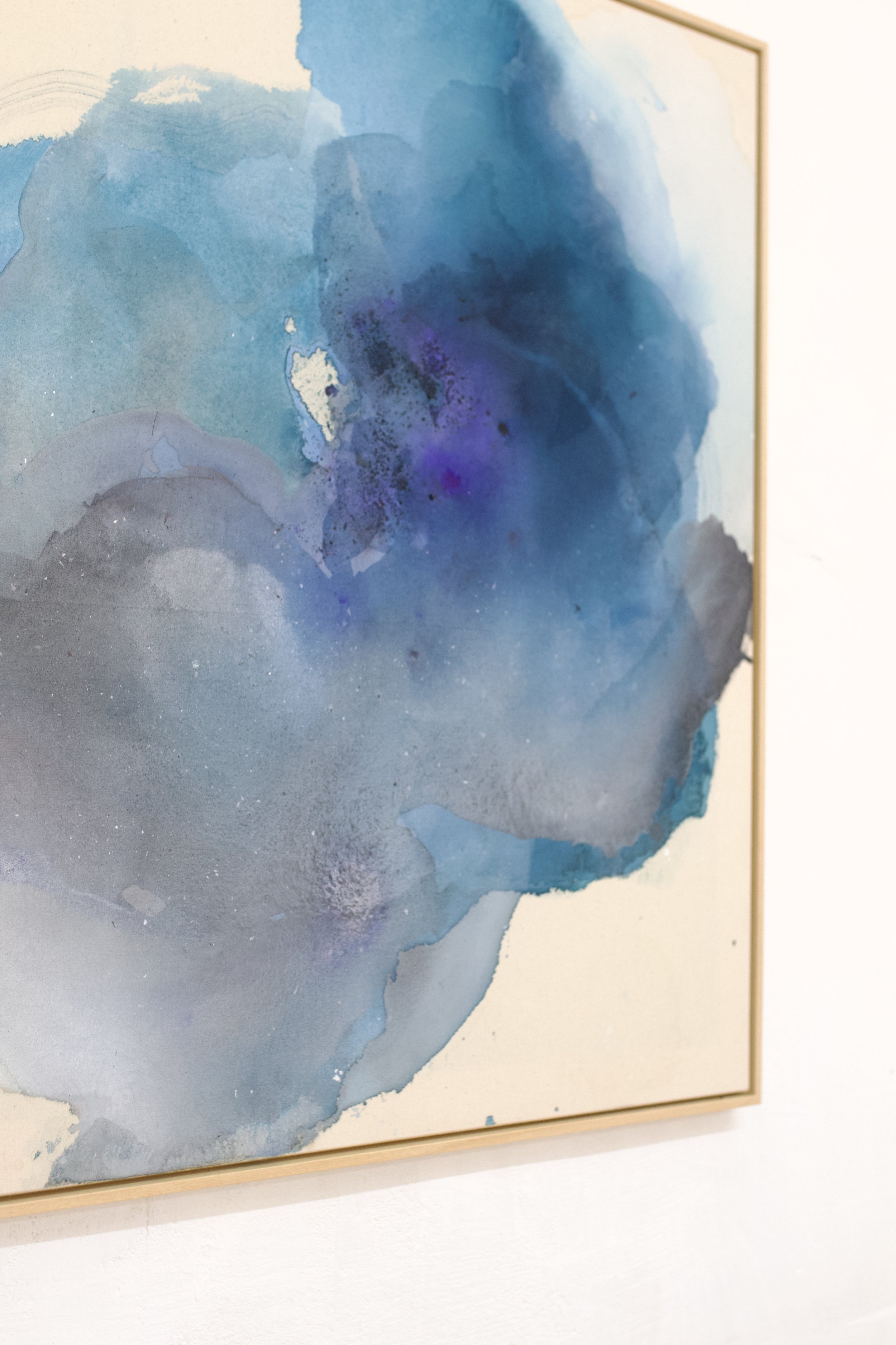 A watercolor painting with shades of blue, purple, and gray. It is framed and mounted on a white wall.
