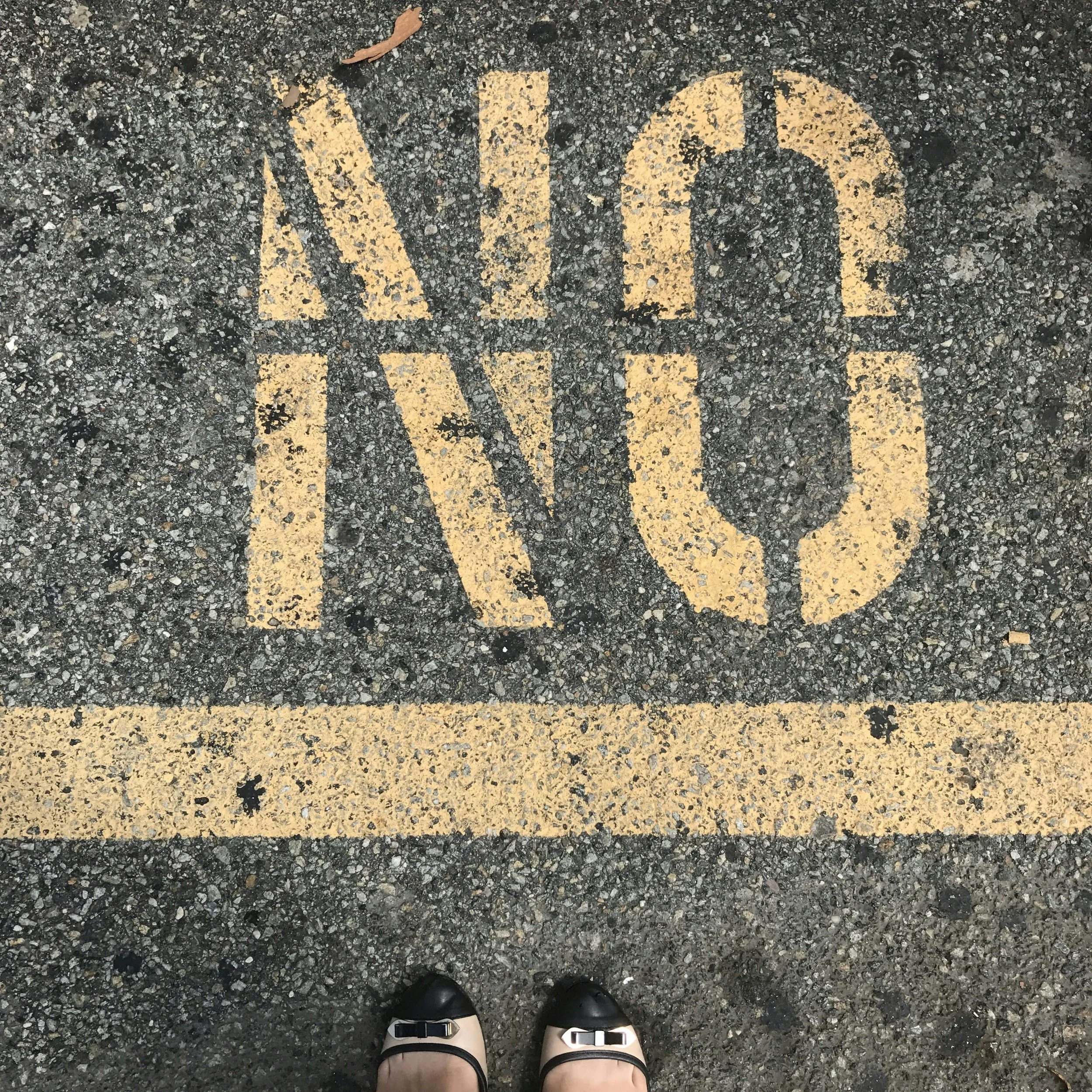 A person's feet wearing black sandals standing on asphalt with yellow road markings reading 'WALK' on a crosswalk.