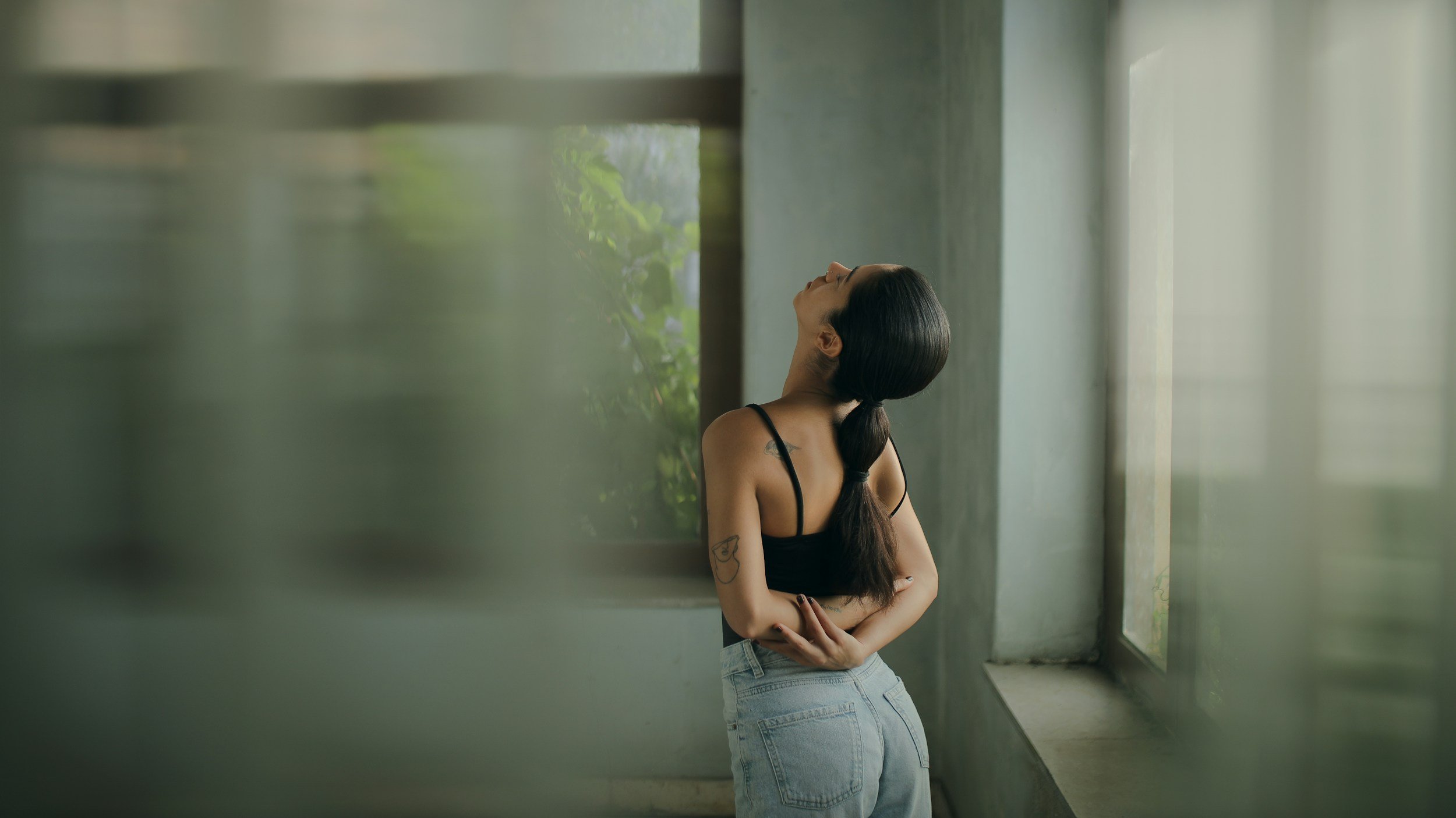 A woman with long dark hair in a ponytail, wearing a black tank top and light blue jeans, stands by a large window with sheer white curtains. She has her eyes closed and appears to be relaxing or enjoying the natural light and greenery outside.