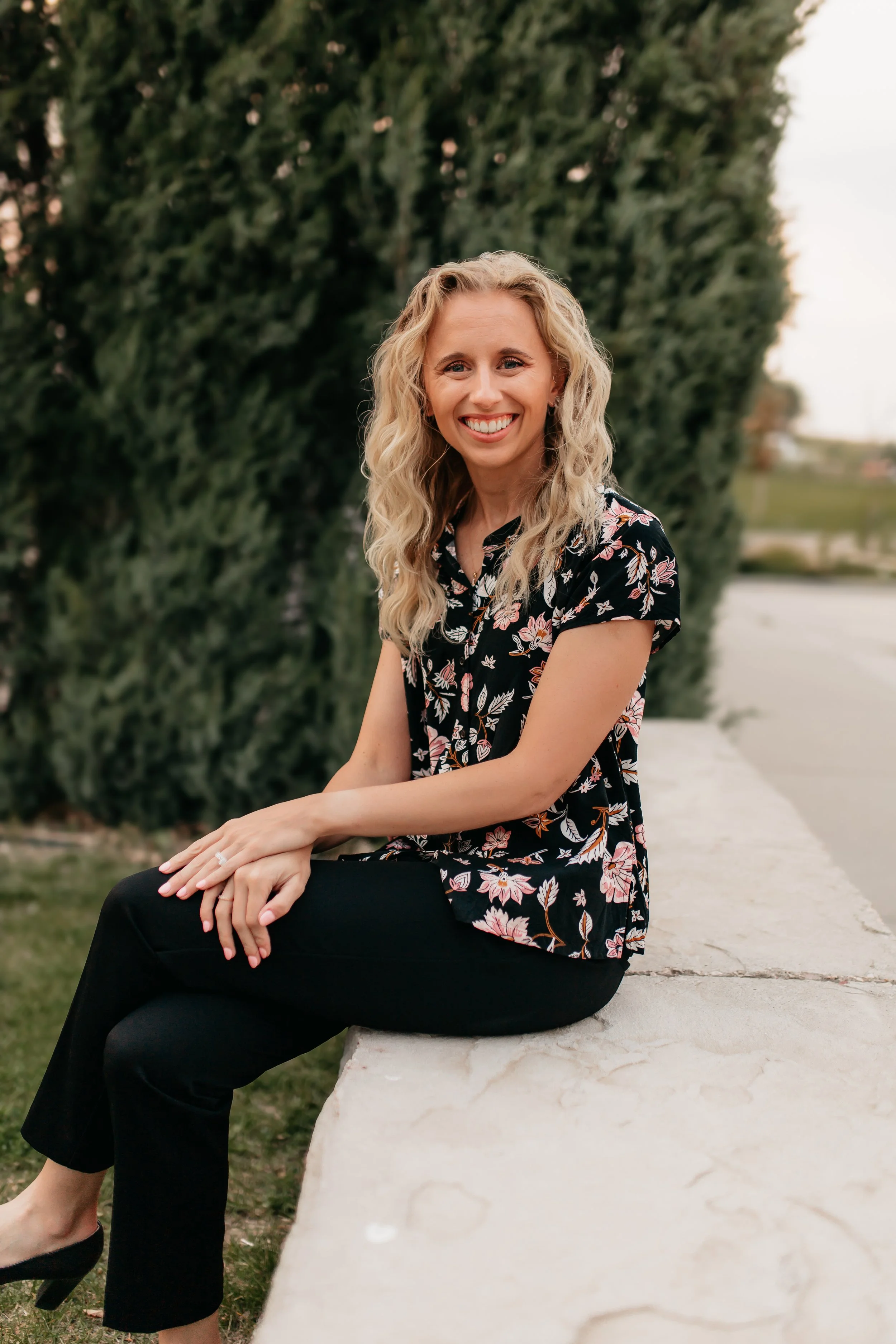 A woman with curly blonde hair smiling and sitting on a stone bench outdoors with green trees in the background.