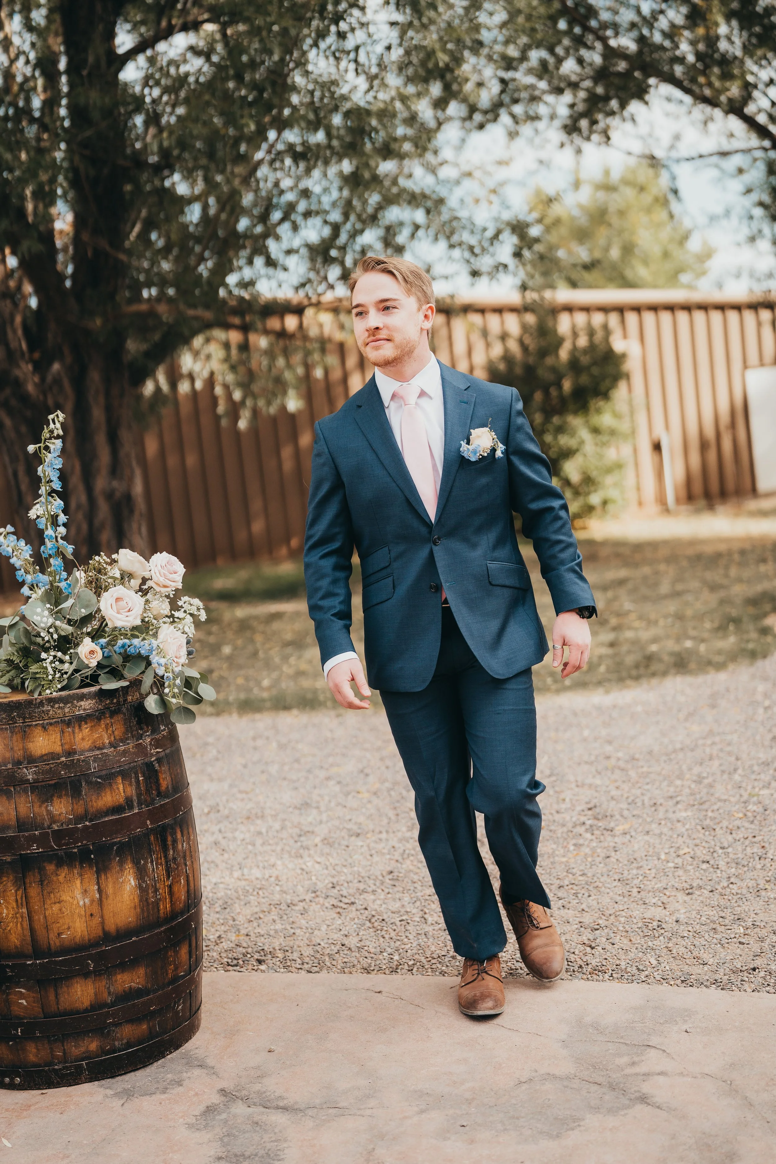 A young man in a blue suit, white shirt, and pink tie walking outdoors near a wooden barrel decorated with flowers on a sunny day.