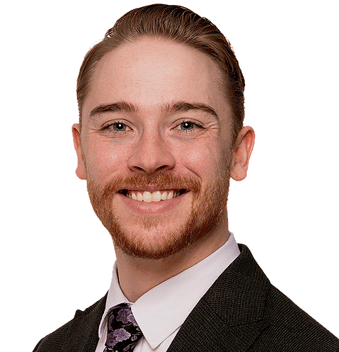 A young man with light skin, smiling, wearing a dark suit, white shirt, patterned tie, and has neatly styled hair and a beard.