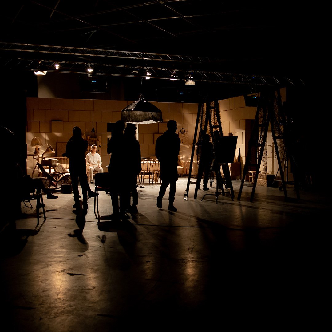 A dimly lit theater stage with silhouettes of people, set decorations, and camera equipment during rehearsal or filming.