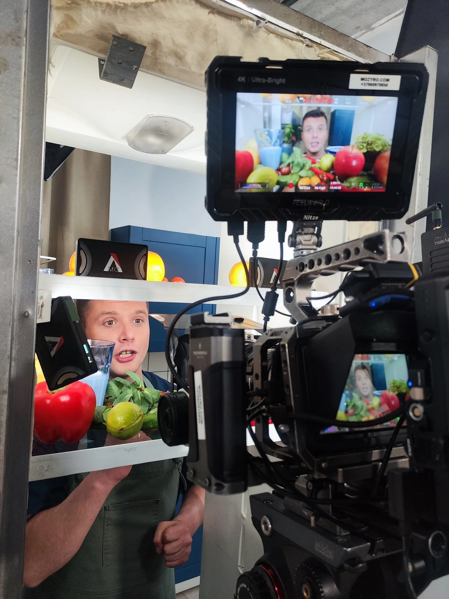 A man in a green apron stands behind a transparent surface with various fruits including red and green apples, oranges, and a lime. He is speaking or explaining something. Behind him, there is a blue cabinet and some yellow lights, and a camera on a rig points at him, capturing the scene for a video or photo shoot.
