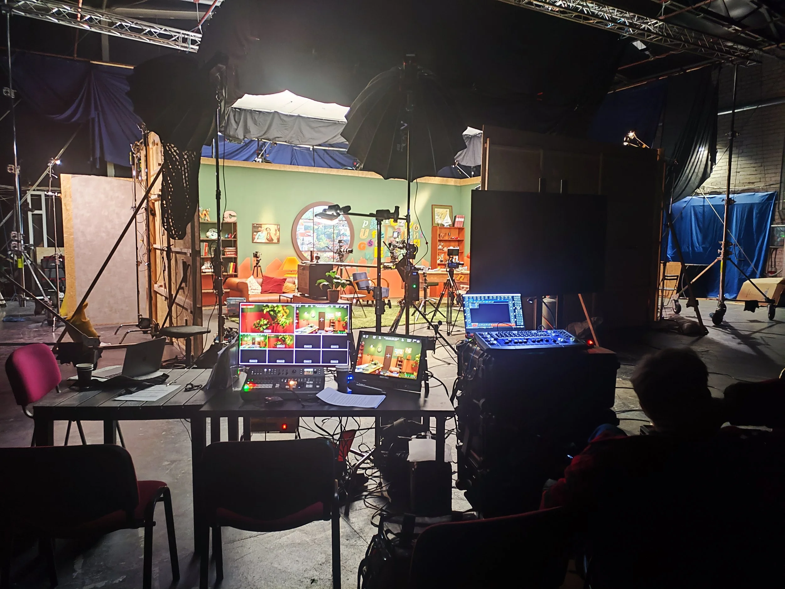 Behind the scenes view of a television or film studio with cameras, lights, and equipment set up for filming, and a cozy living room set with a green wall, bookshelf, and decorations.