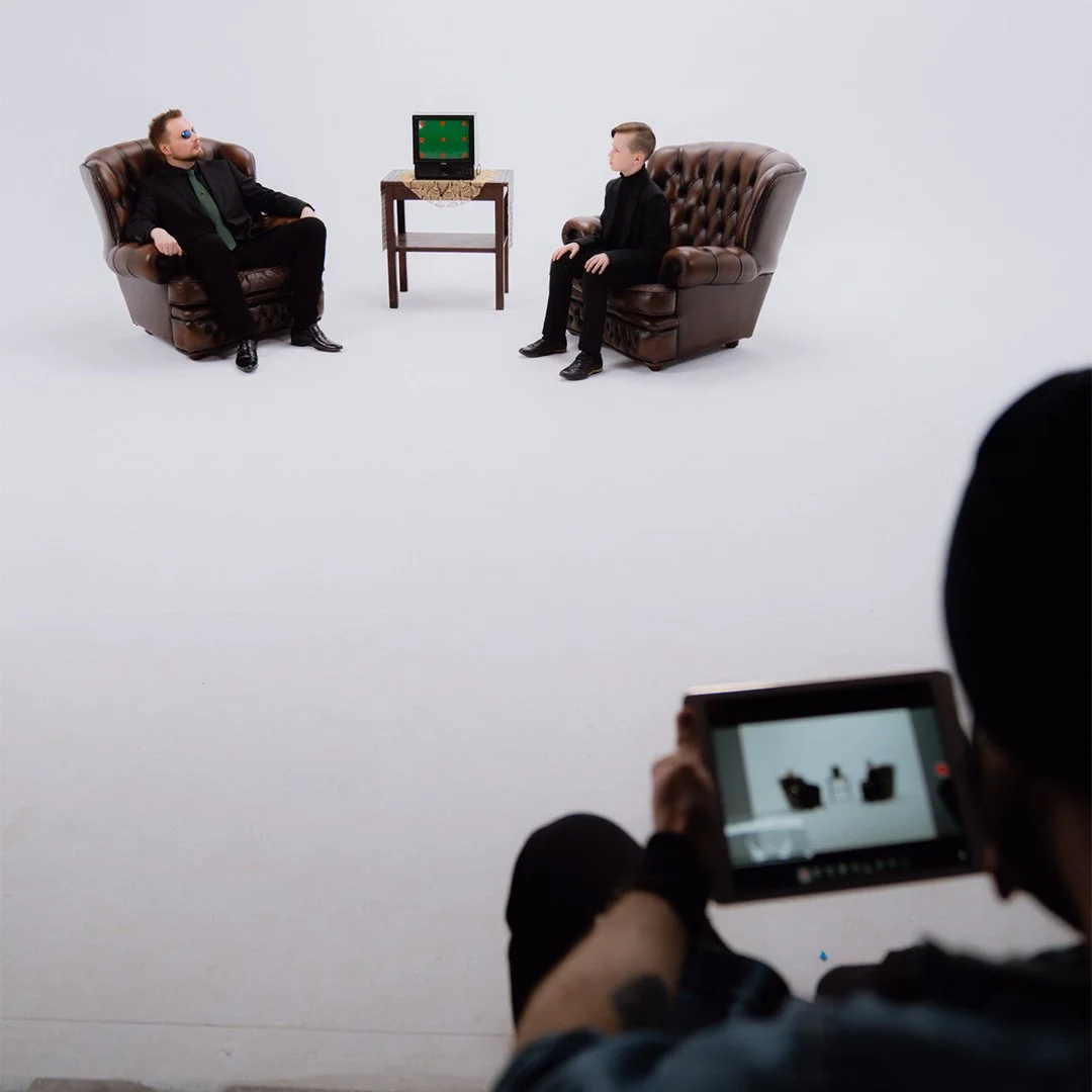 Two men sitting on leather armchairs facing each other, with a small table and a vintage television between them, and a person in the foreground filming the scene with a tablet.