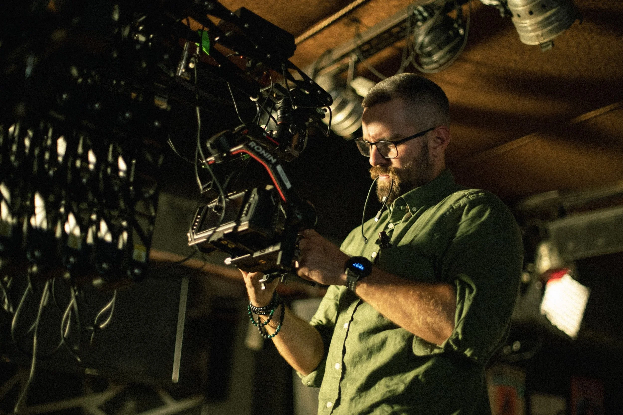 A man with glasses and a beard inspecting equipment in a dimly lit room with a wooden ceiling, surrounded by technical gear and cables.
