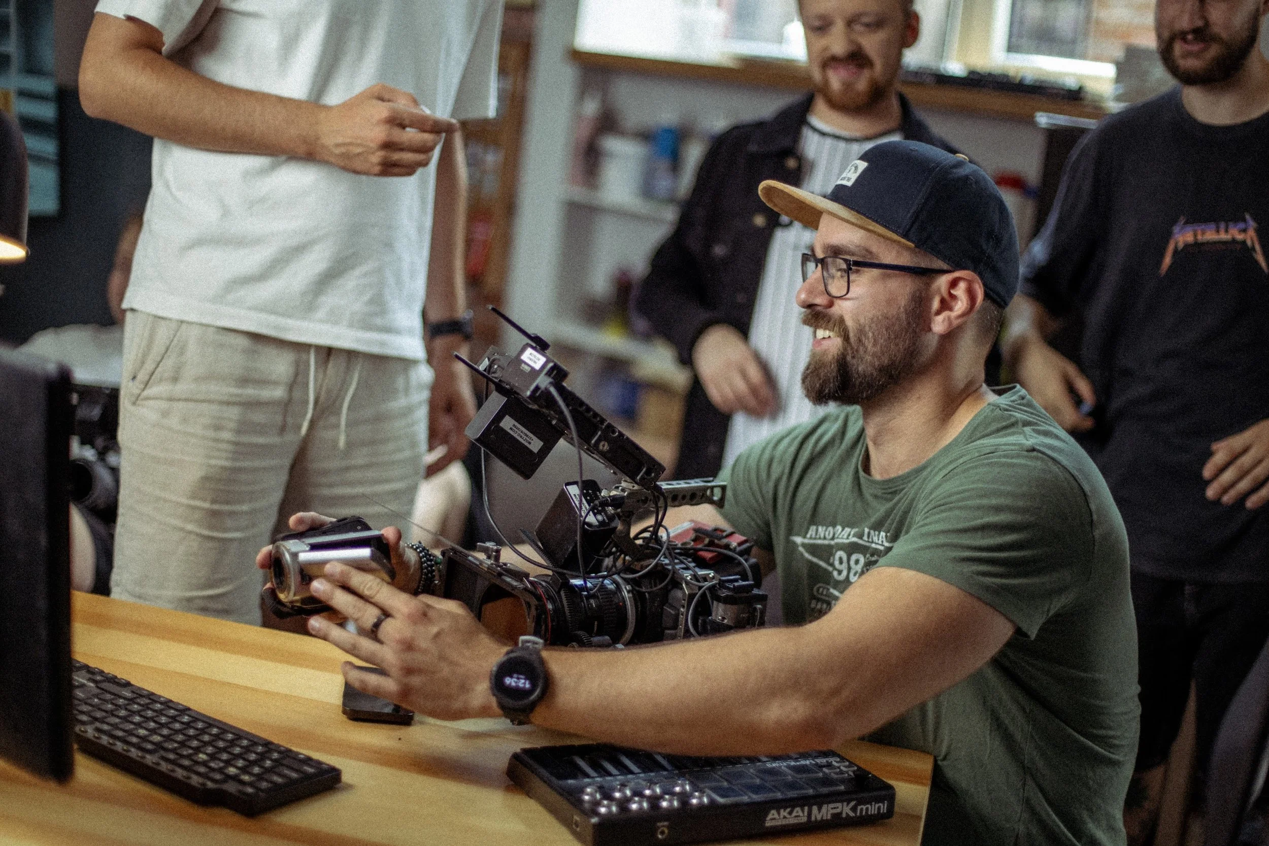 A group of people in a room with a wooden table, working on electronic or digital devices, with one person holding a camera and others standing or sitting around the table, engaged in a discussion or collaboration.