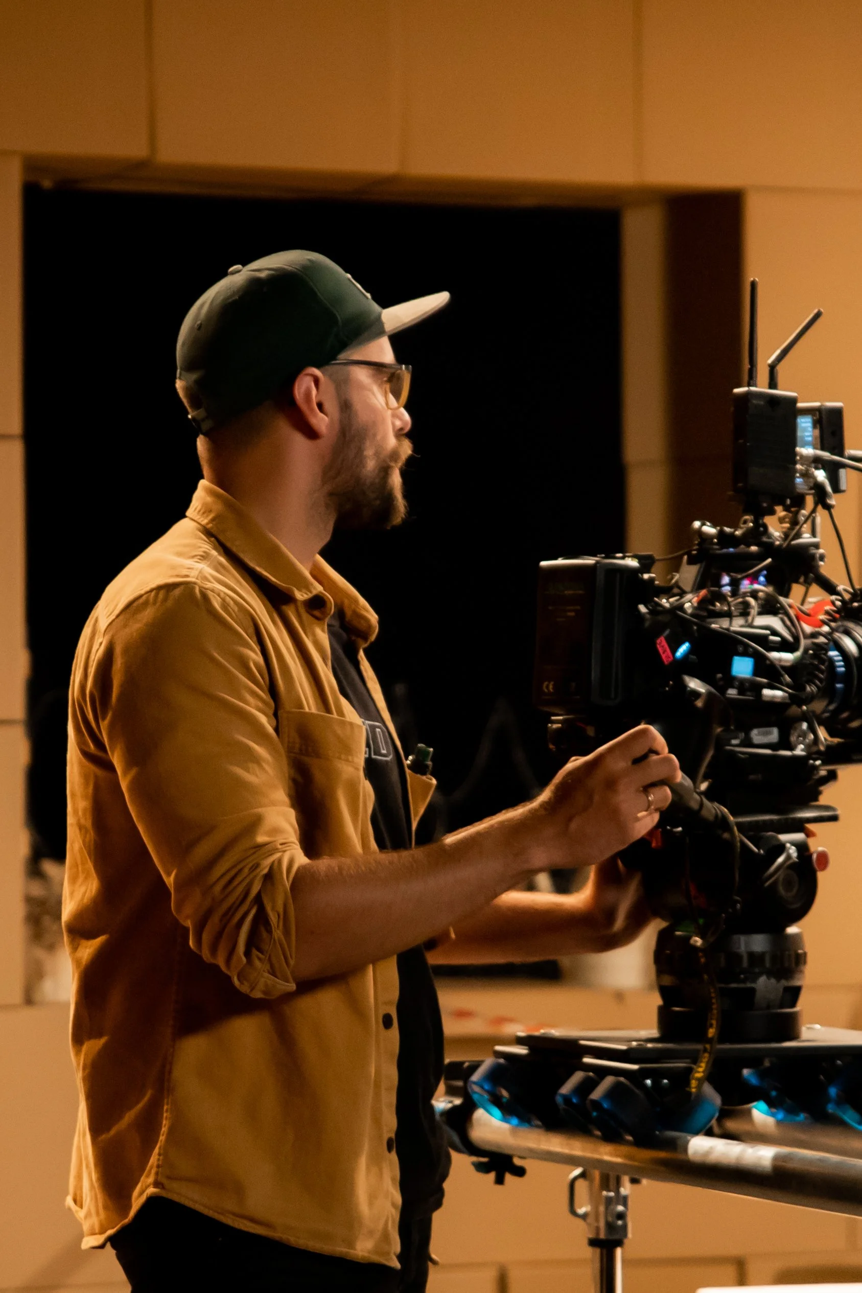 A man wearing glasses and a baseball cap operating a professional camera on a tripod in a dimly lit room.