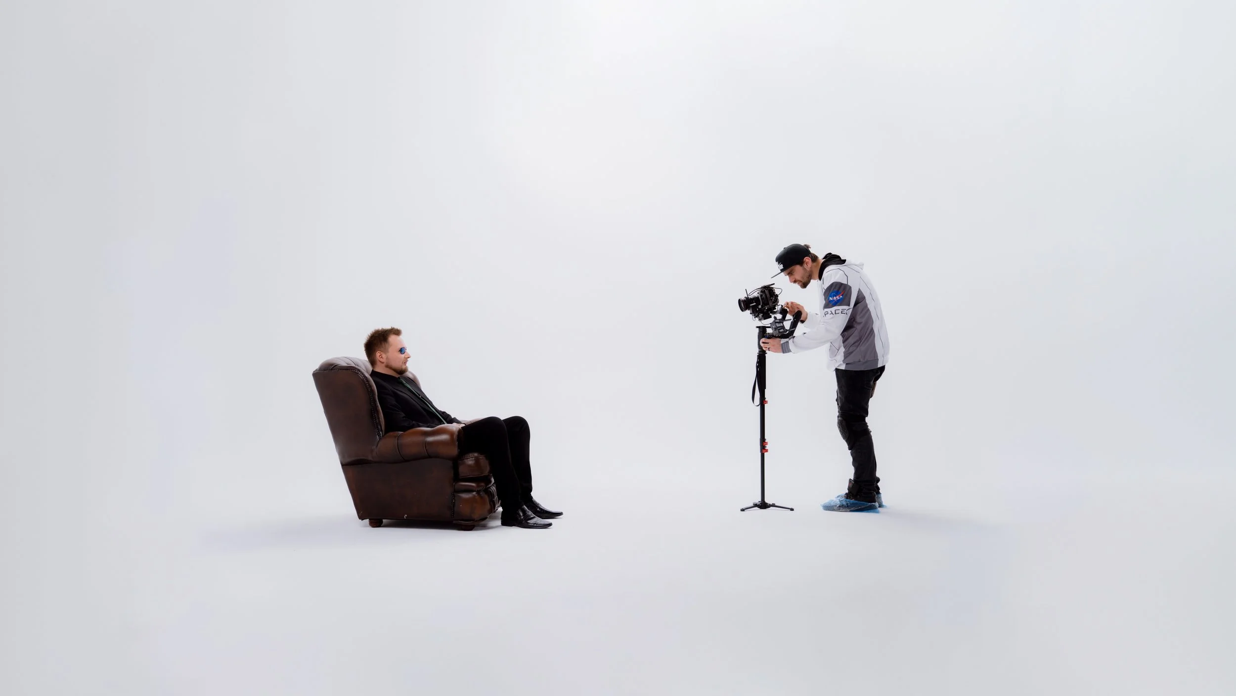 A man sitting in a leather armchair with sunglasses while a cameraman films him against a plain white background.