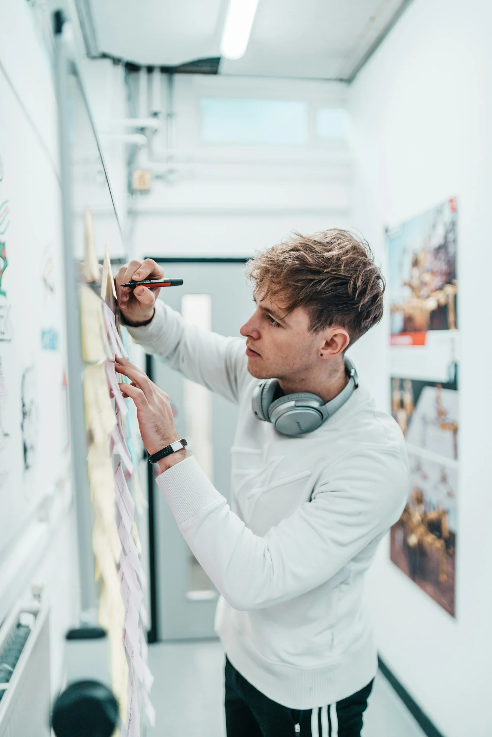 A young man with curly hair and headphones around his neck is writing on a sticky note and placing it on a board with other notes in a bright hallway.