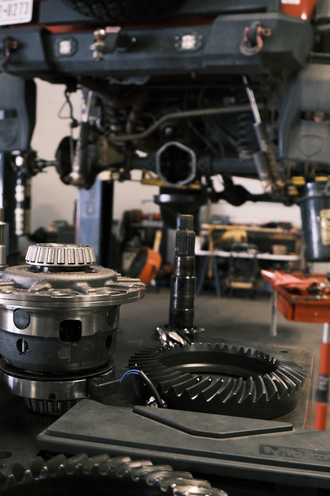 Close-up view of mechanical gears, bearings, and differential components, with a partially assembled transmission in the background, in a workshop setting.