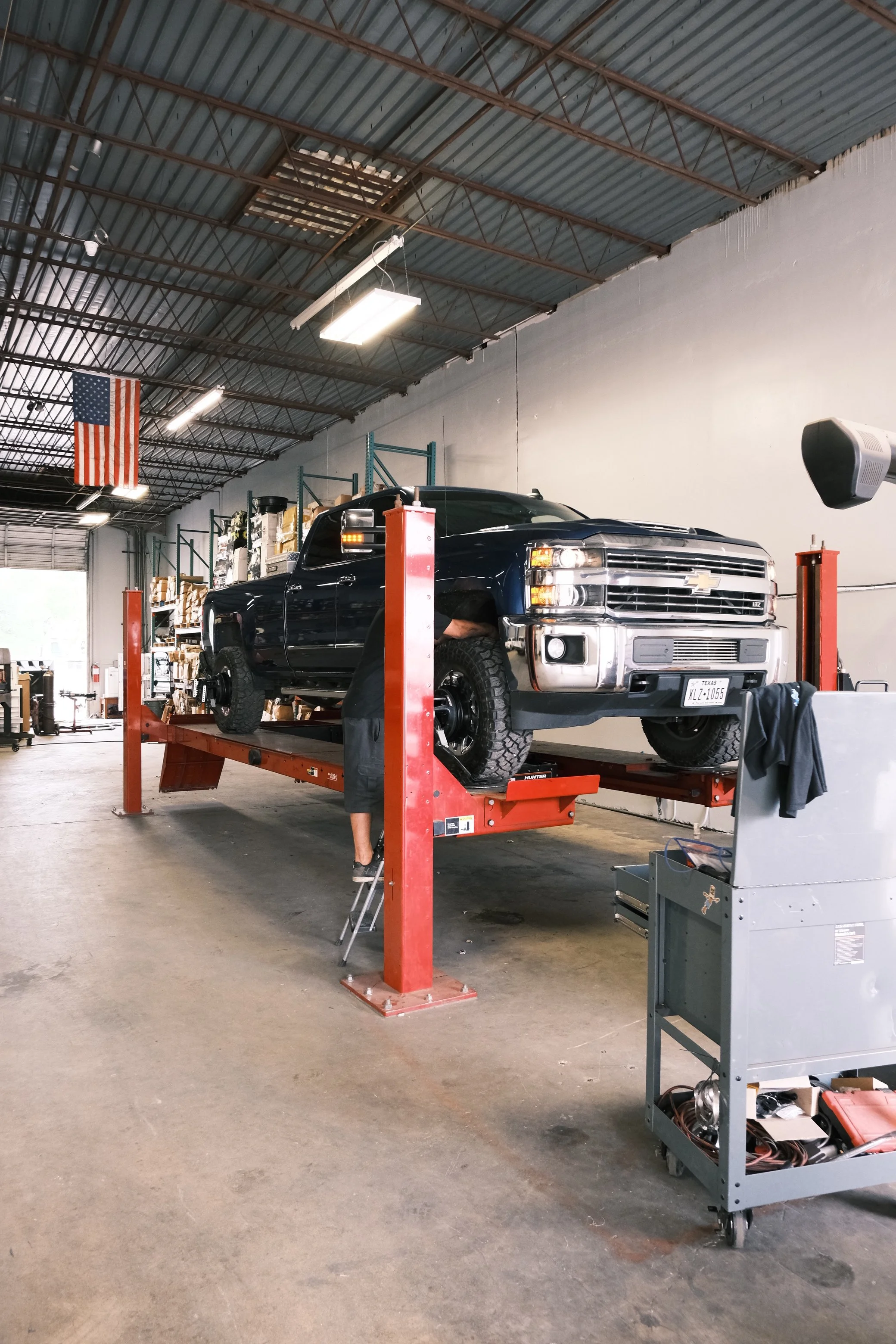 A black Chevrolet Silverado pickup truck is elevated on a hydraulic lift inside a garage or auto repair shop. The shop has a high, industrial ceiling with exposed metal beams, fluorescent lighting, and an American flag hanging from the ceiling. Shelves with various items are visible in the background.