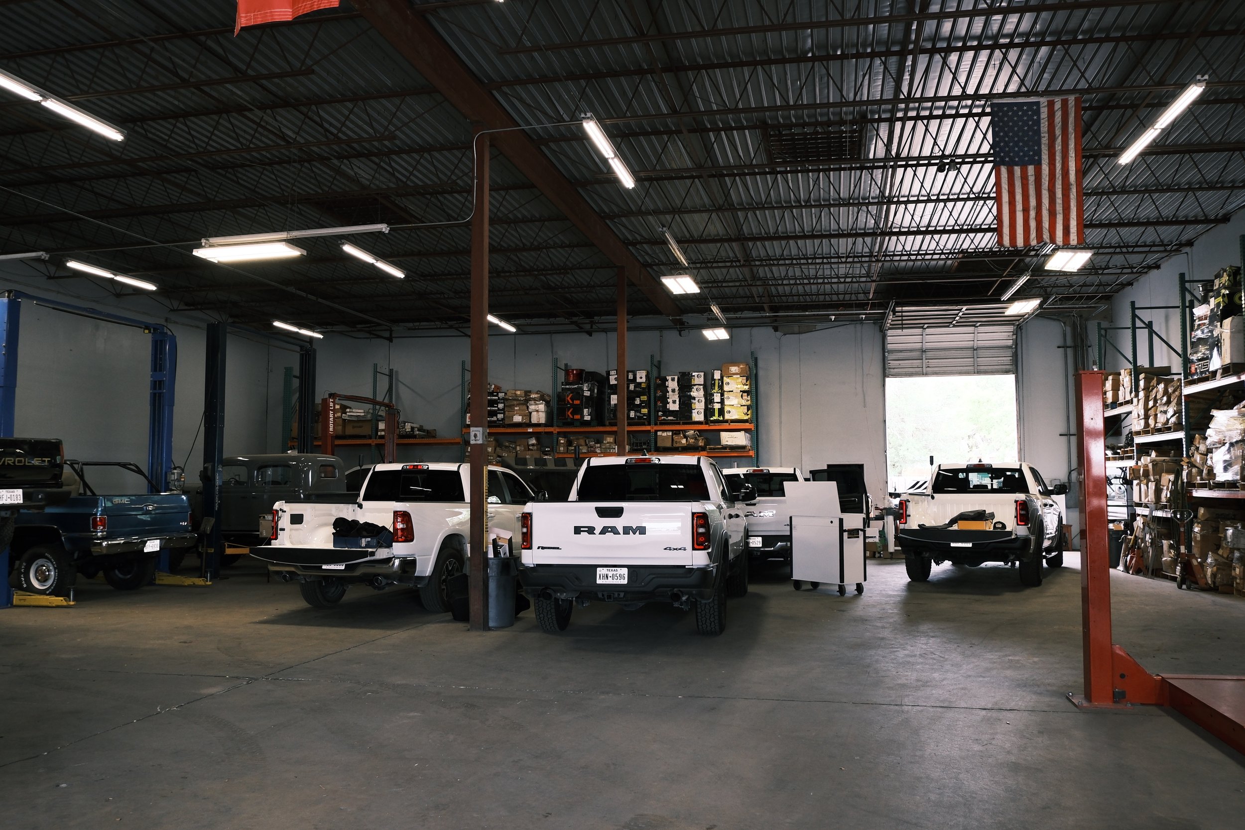Inside a warehouse with several parked trucks, metal shelves filled with boxes and supplies, and an American flag hanging from the ceiling.