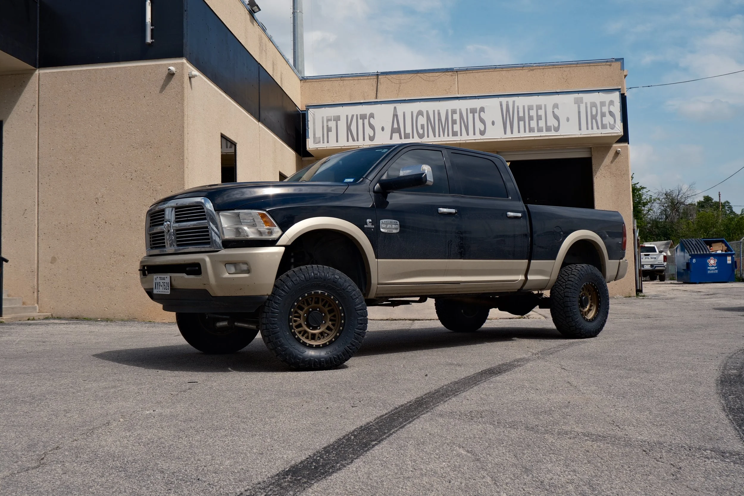 A black and beige lifted Dodge Ram truck with large tires parked in front of an automotive repair shop. The shop has a sign that reads 'Lift Kits, Alignments, Wheels, Tires'.
