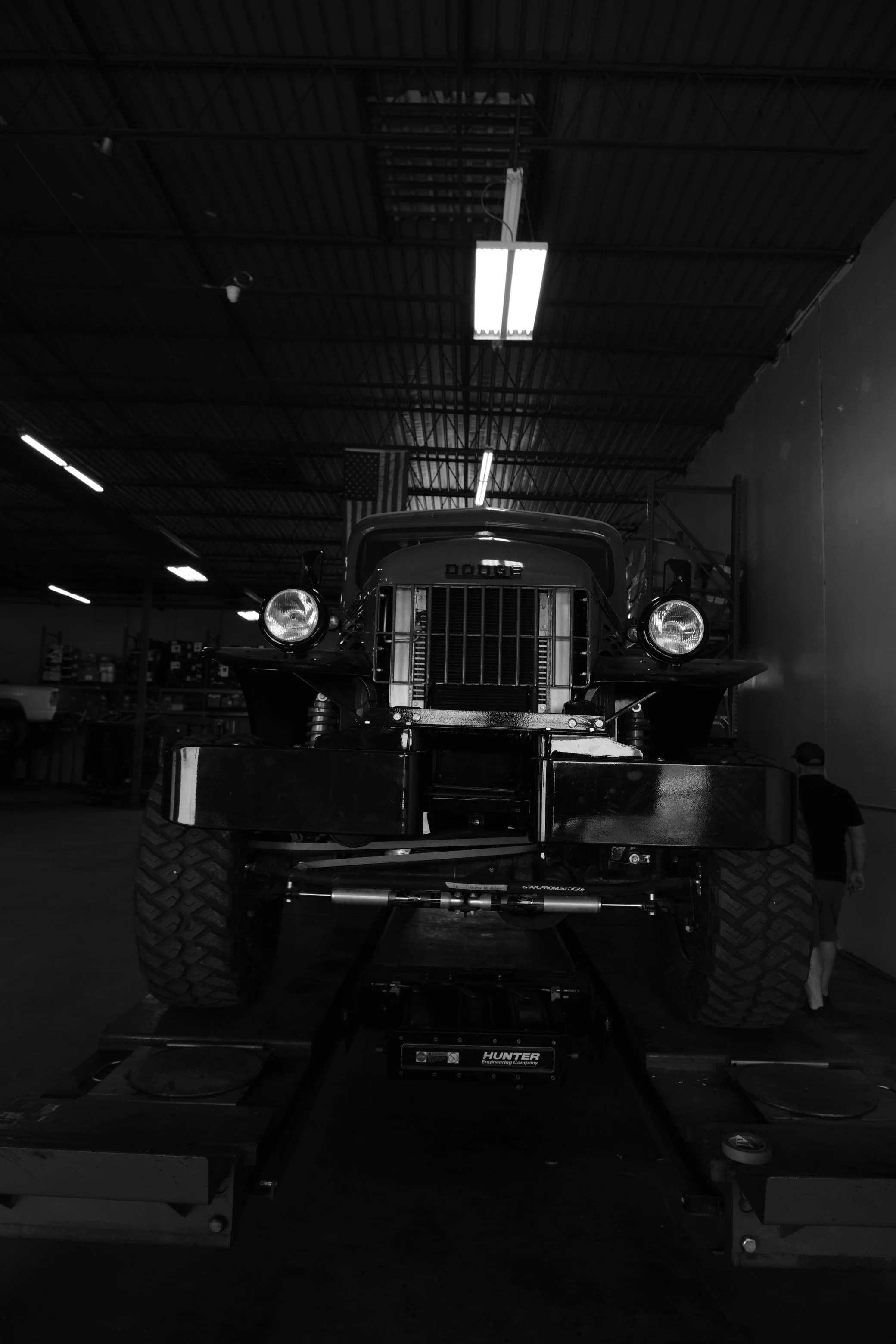 Black and white photo of a vintage Dodge truck in a warehouse or garage with a high ceiling, fluorescent lights, and an American flag hanging in the background.