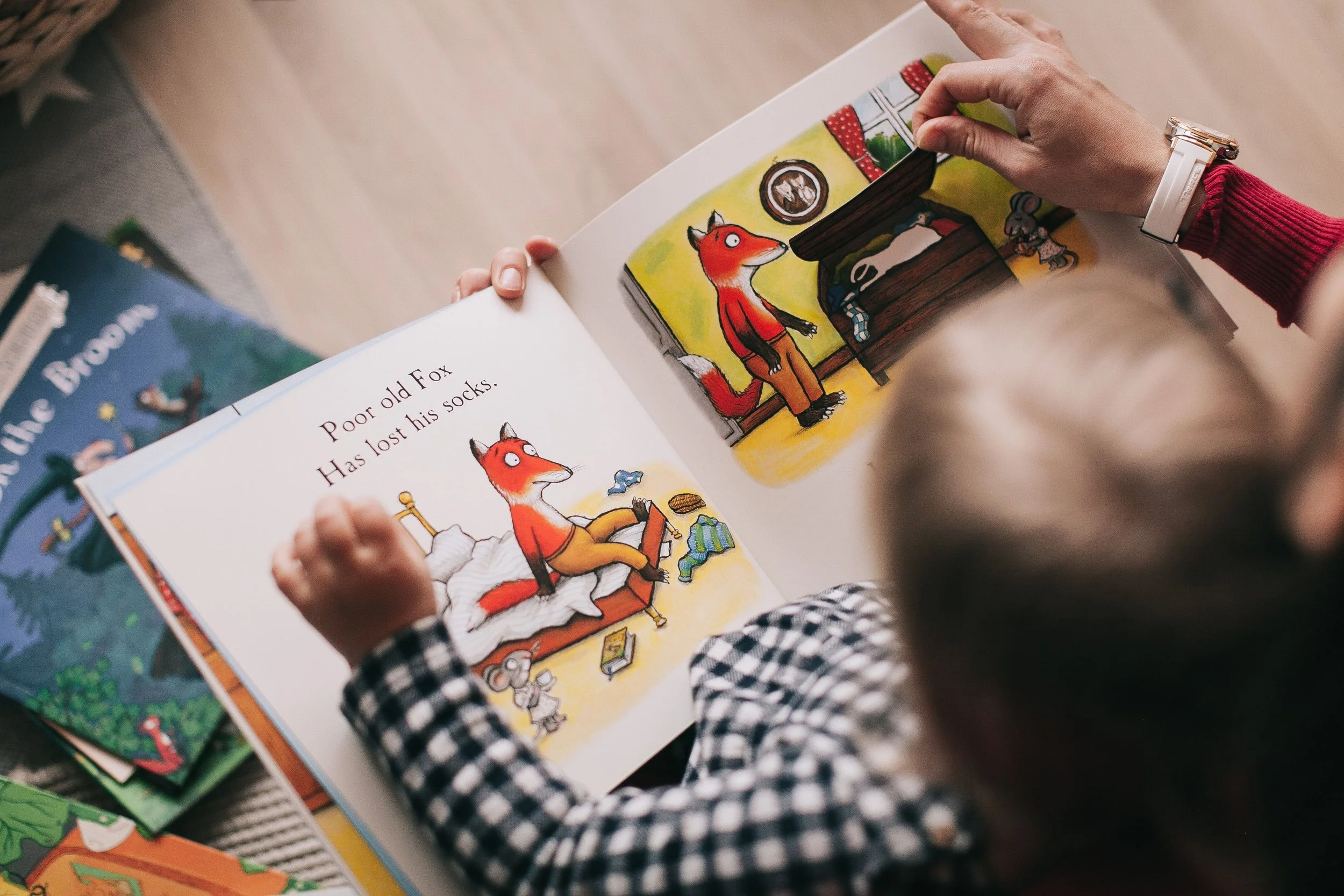 A person with a checkered shirt and white wristwatch reading a colorful illustrated children's book with animal characters, including a fox, on a wooden floor with other children's books nearby.