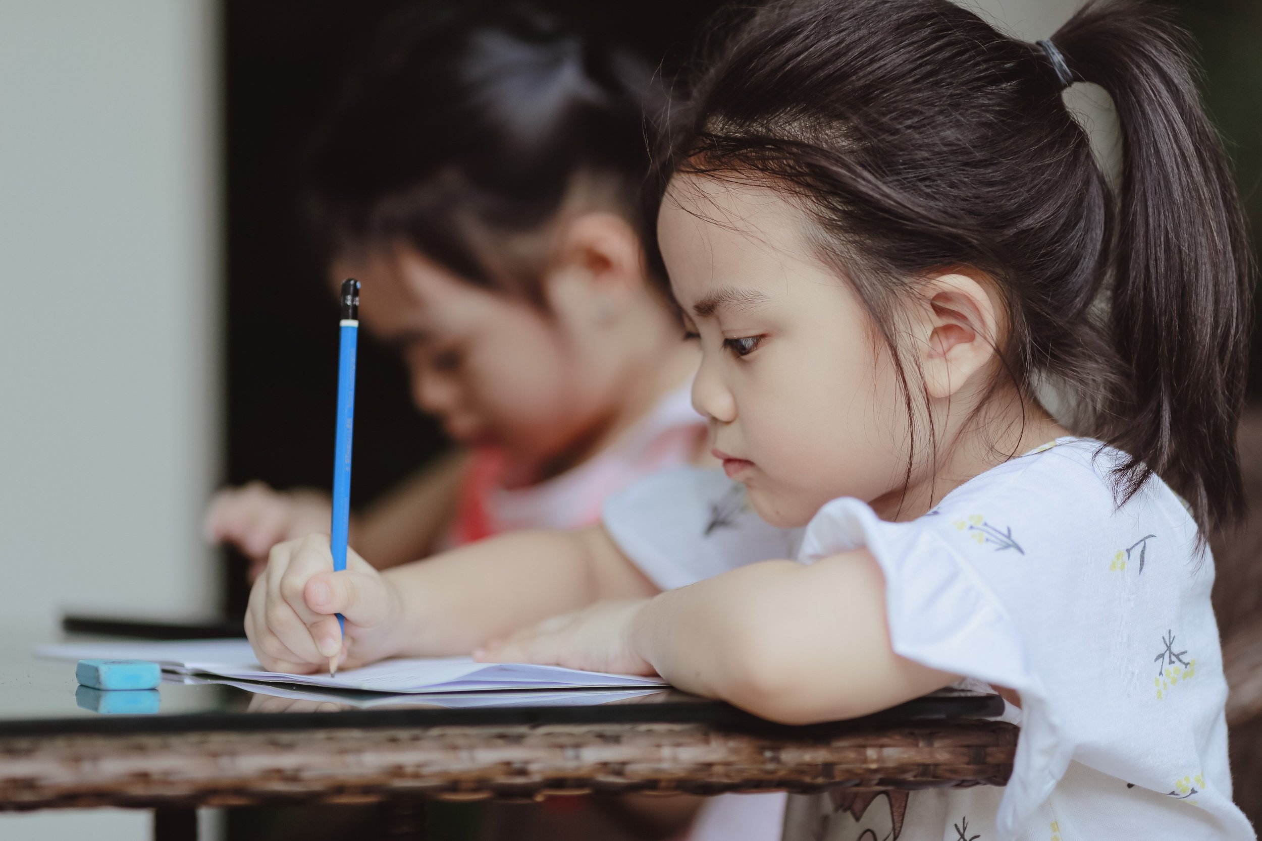 Two young children, a boy and a girl, are sitting at a table, focused on writing or drawing on paper with a blue pen. The girl has long brown hair tied back and is wearing a white shirt with small colorful prints. The boy has dark hair and is wearing glasses.