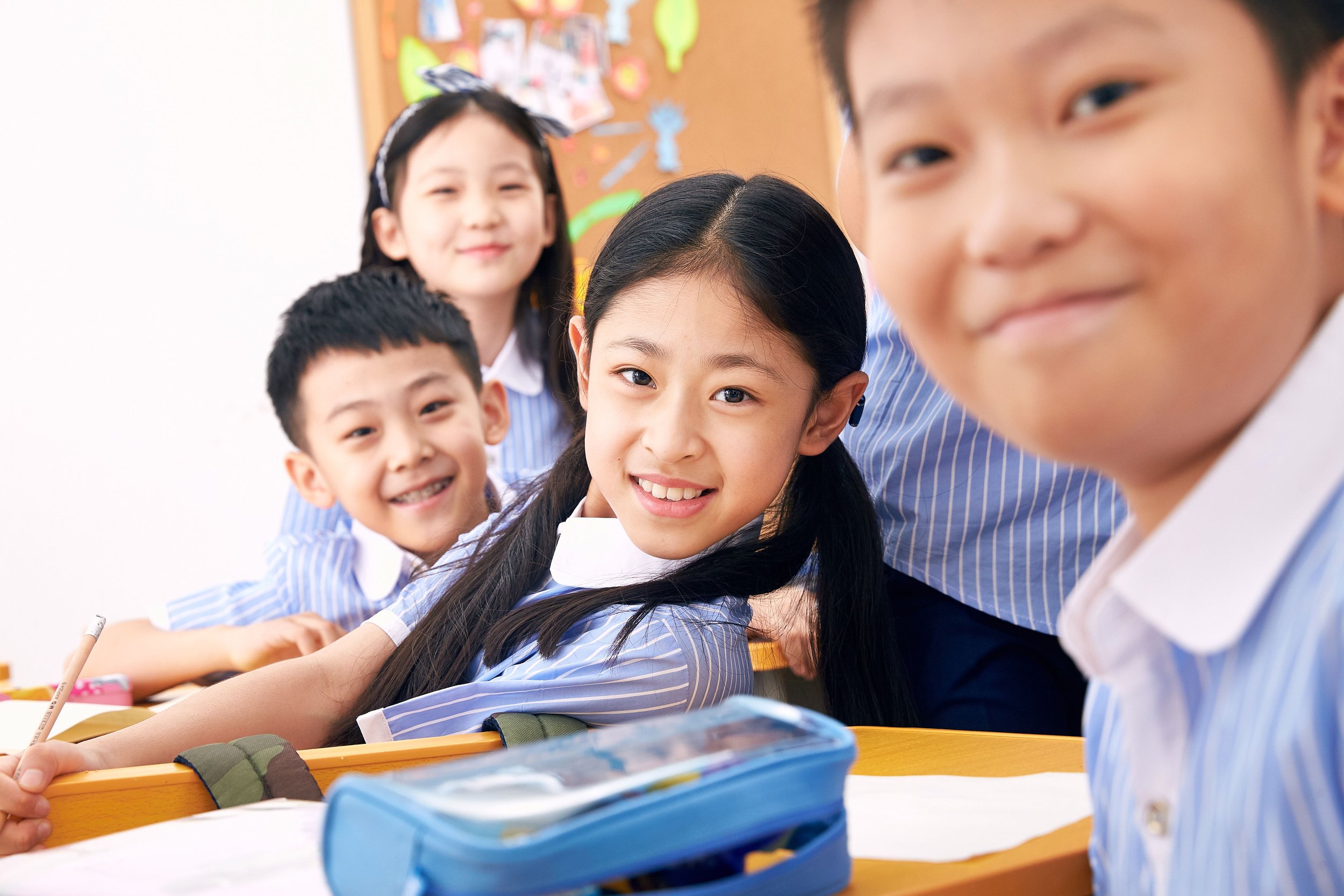 Smiling elementary school students in blue uniforms sitting at desks in a classroom, with some students leaning forward and looking at the camera.