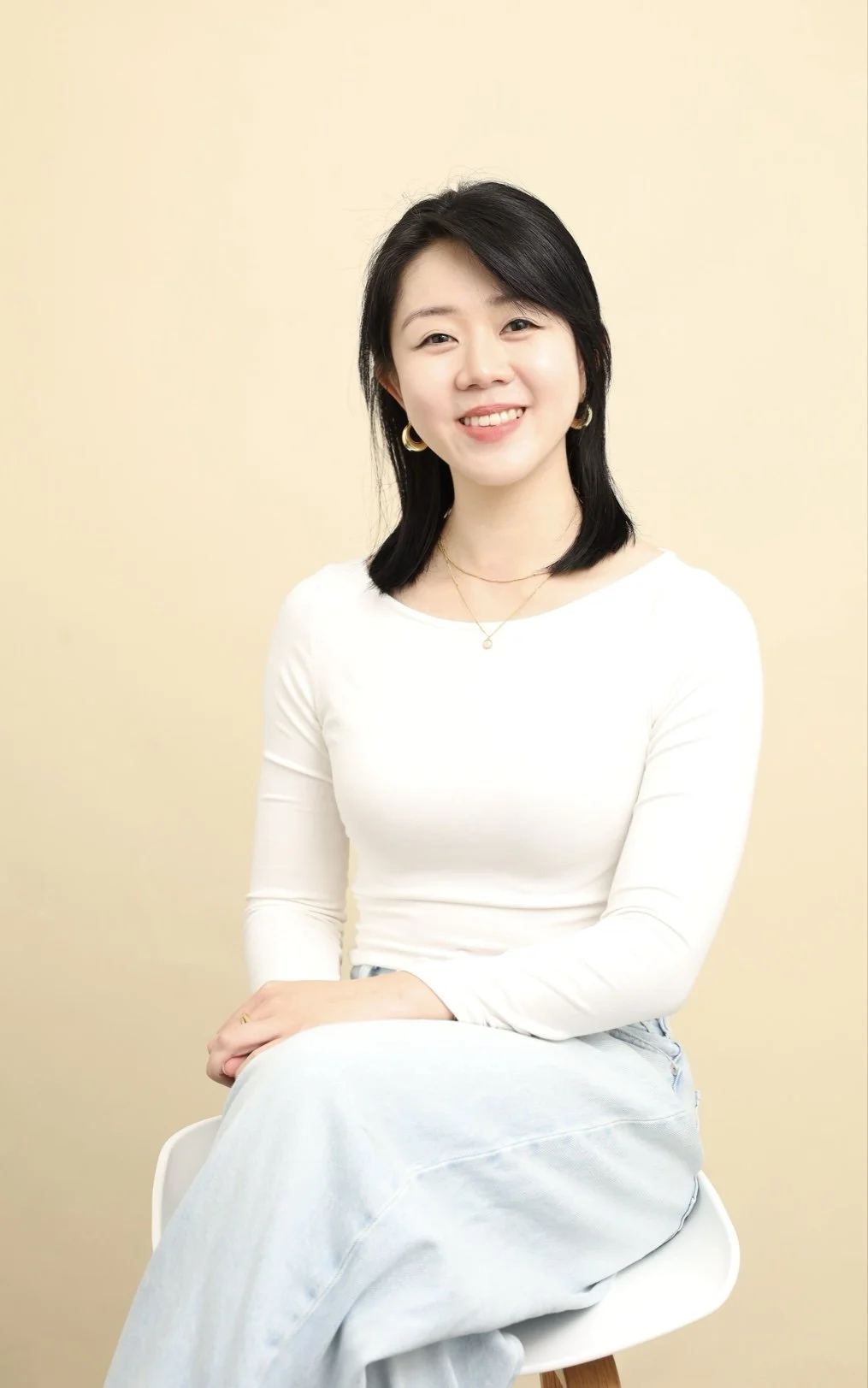 A young woman with black hair, wearing a white long-sleeve top, light blue jeans, and gold jewelry, sitting on a white chair against a beige background, smiling at the camera.