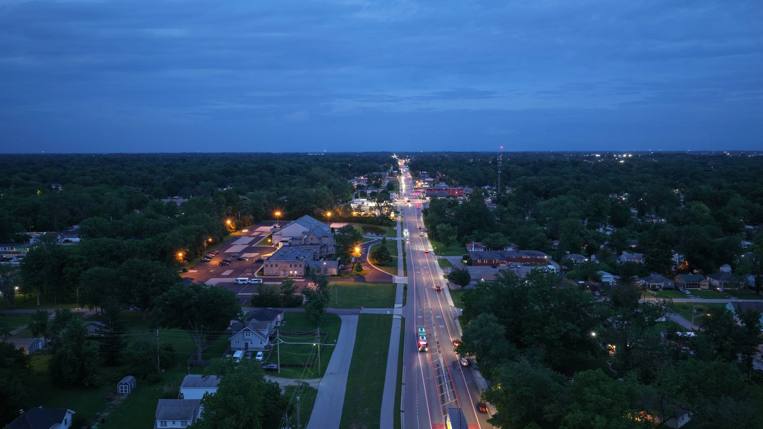 Aerial view of a small town at dusk, showing a main road with traffic, residential houses, and green trees, under a blue sky with clouds.
