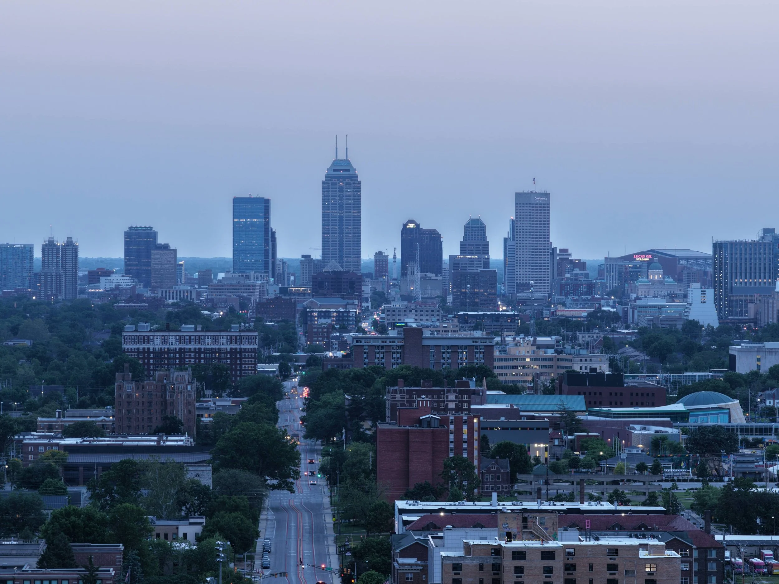 A city skyline with tall buildings, including the Empire State Building, during early evening with a cloudy sky and city lights beginning to turn on.