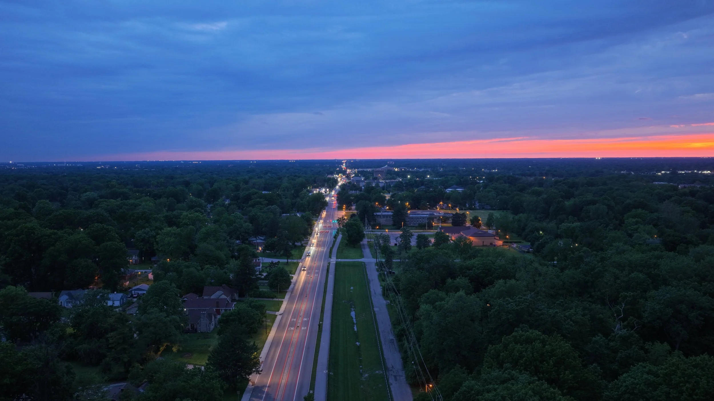 Aerial view of a town at dusk with a sunset sky, a main road running through a green, wooded area, and city lights beginning to turn on.