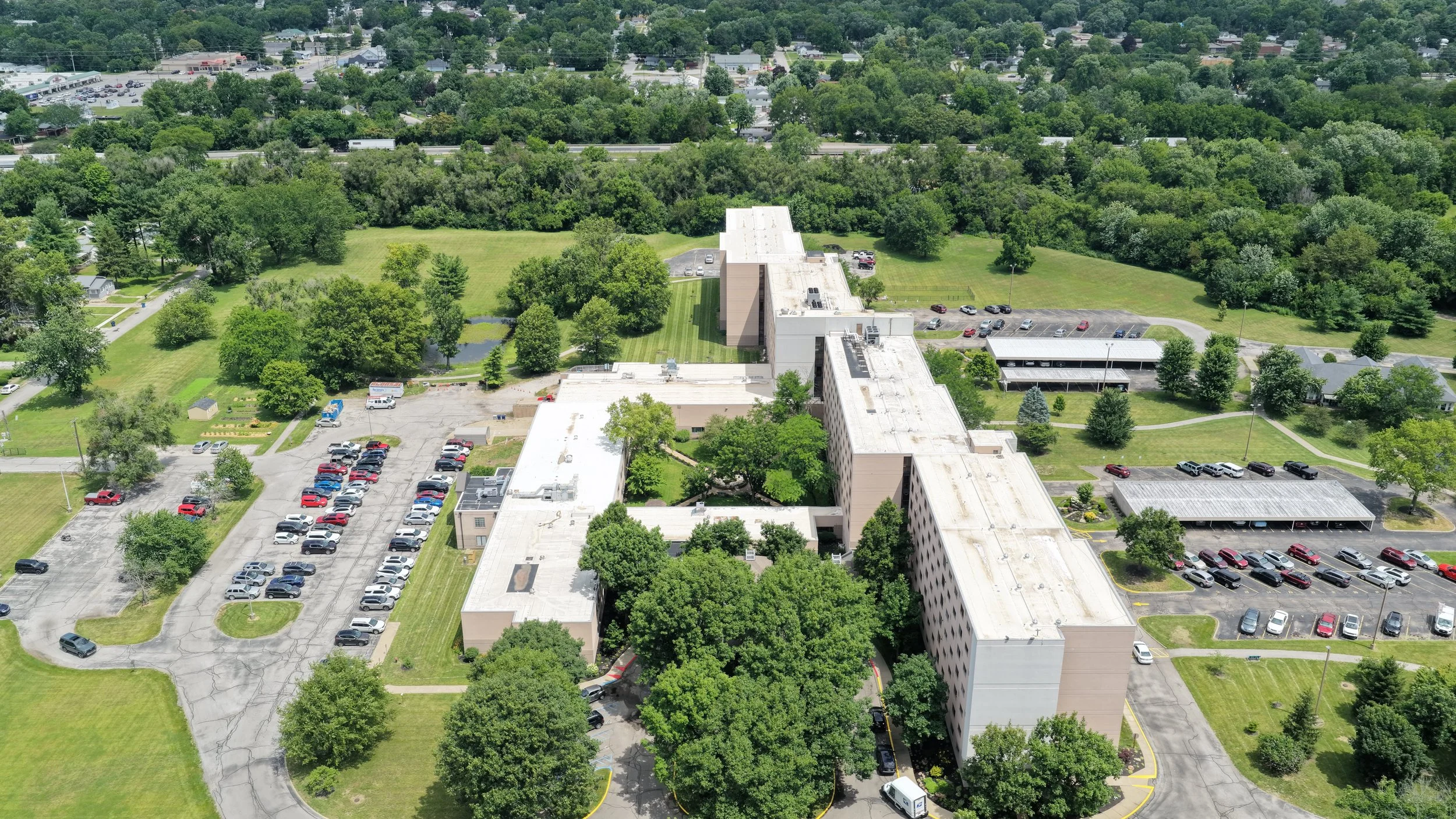 Aerial view of a large building complex surrounded by parking lots, green lawns, trees, and a wooded area in the background.
