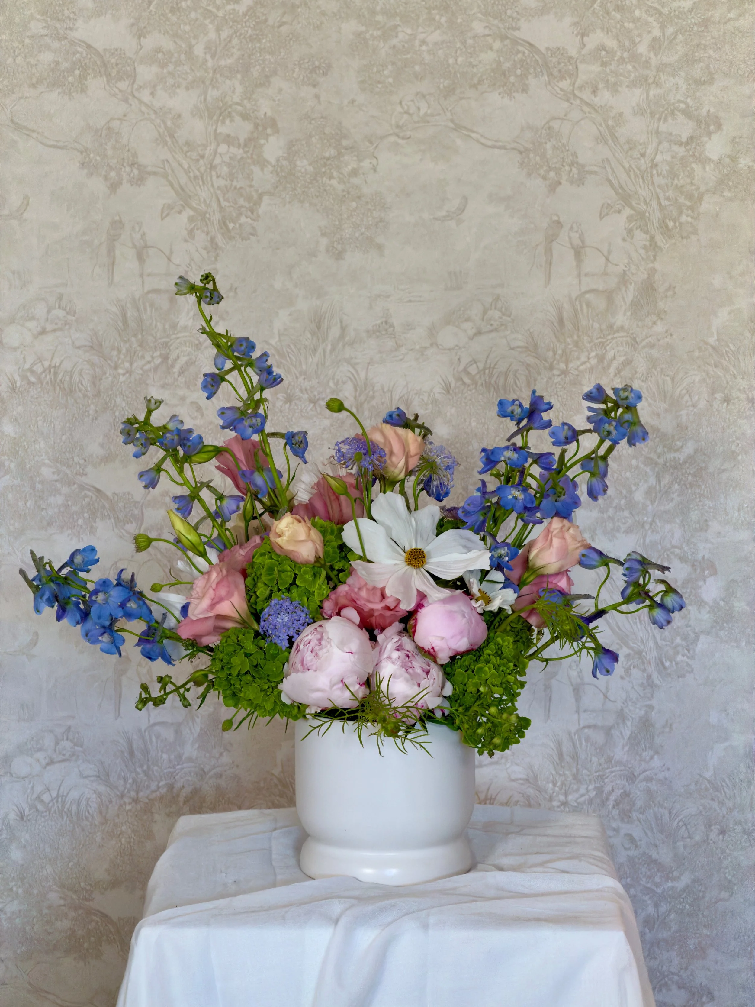 A flower arrangement with pink roses, white lilies, blue delphiniums, and green hydrangeas in a white ceramic vase on a white cloth on a table, with a beige textured wall in the background.