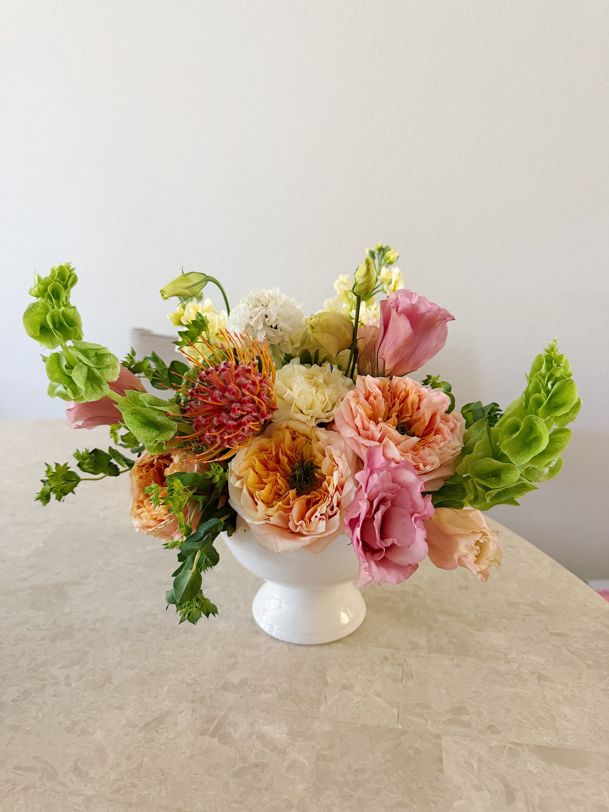 A white vase with a bouquet of pink, white, cream, and orange flowers on a beige marble surface against a plain white wall.