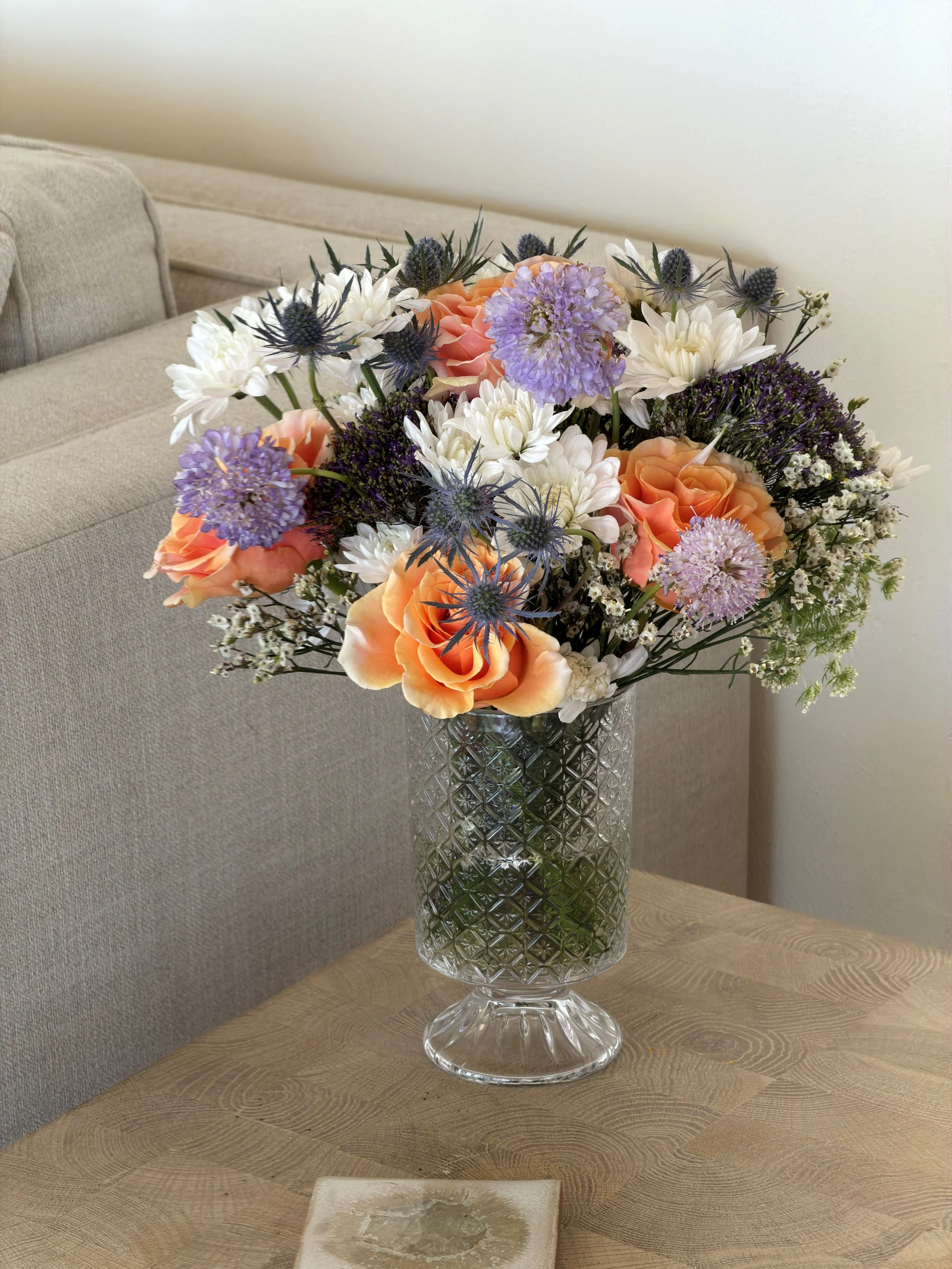 A colorful floral arrangement in a tall, textured glass vase on a wooden table, with a beige sofa and wall in the background.