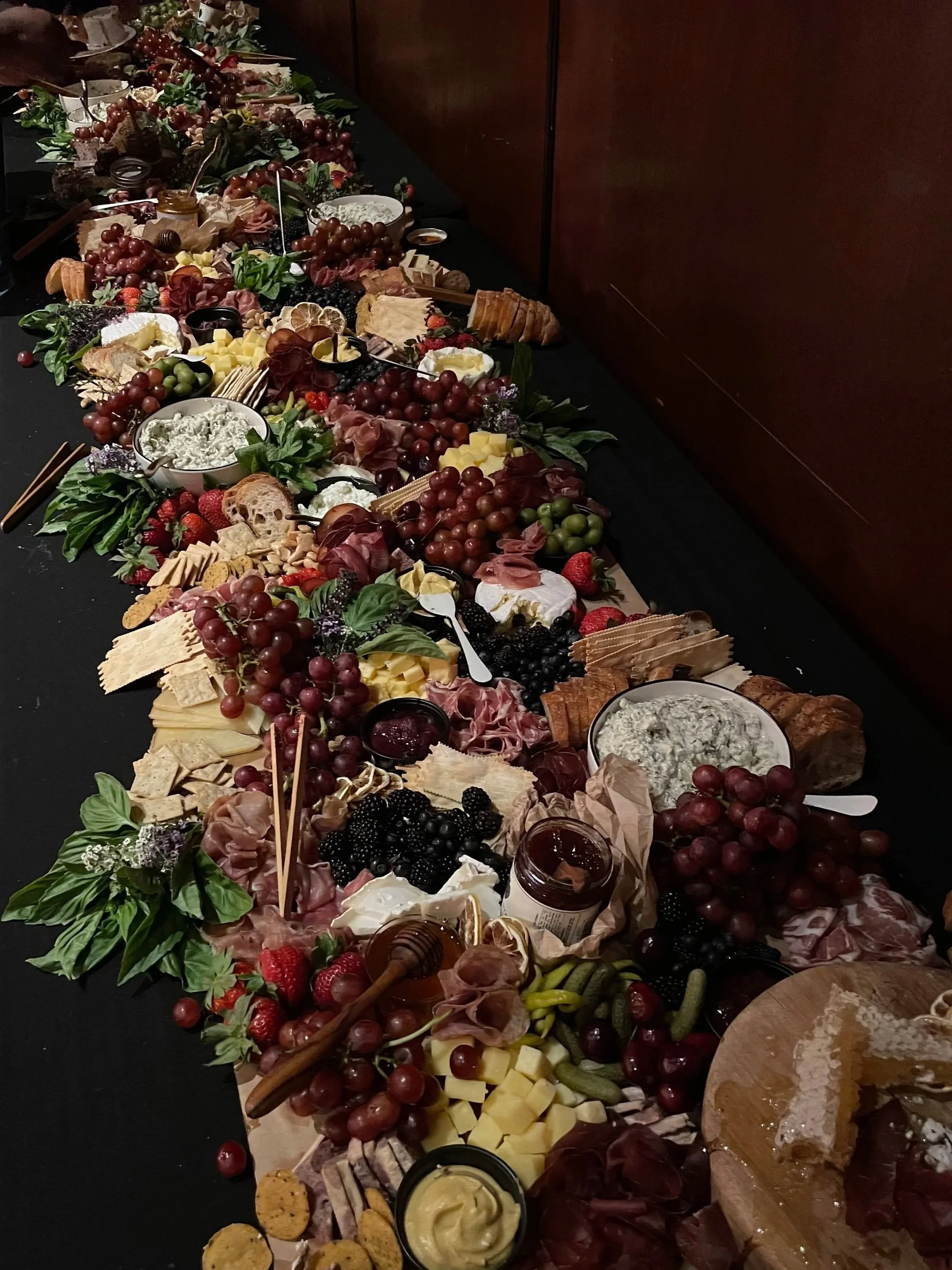 A long table with a black tablecloth, filled with various cheeses, grapes, strawberries, meats, crackers, bread, dips, and other finger foods, decorated with greenery and flowers.