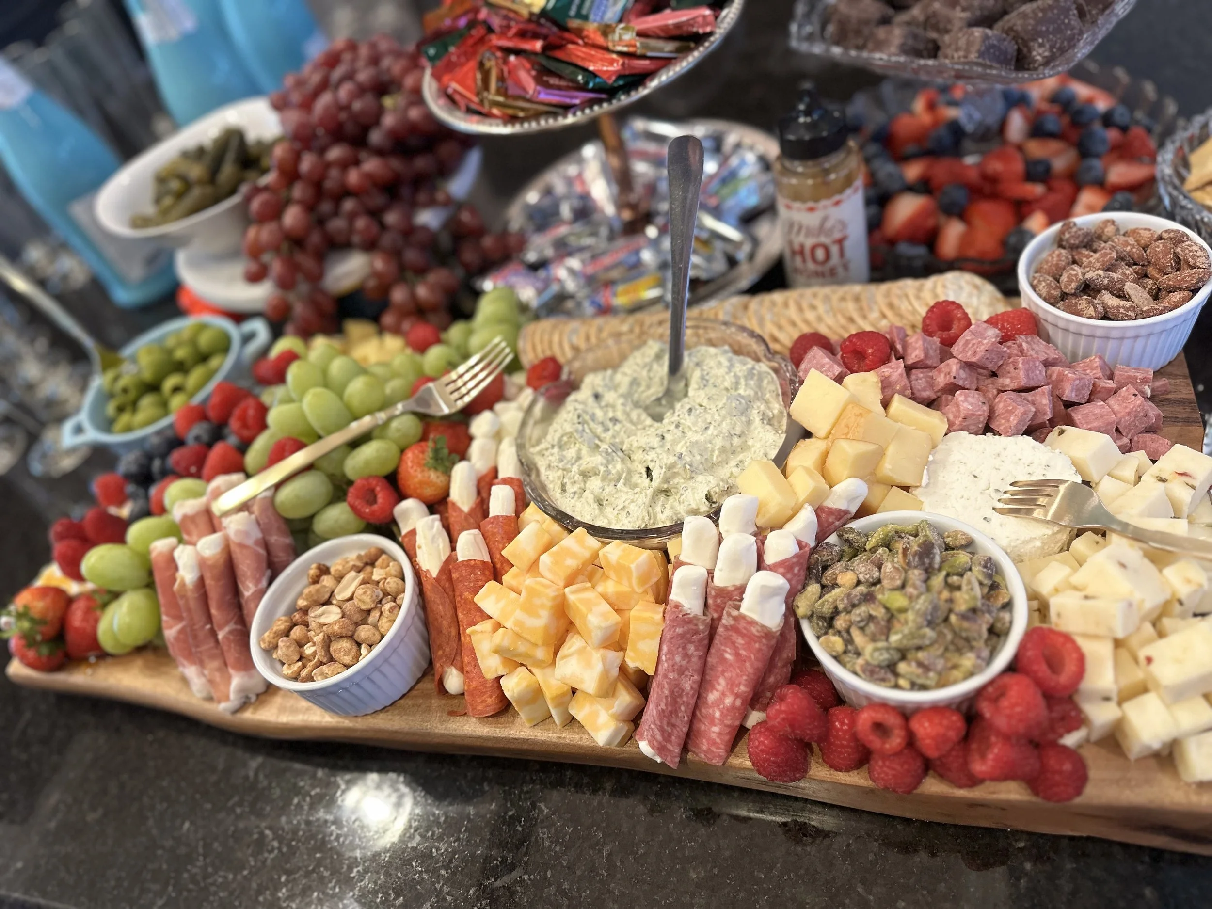 A platter of assorted cheeses, fruits, and snacks including grapes, strawberries, raspberries, cubed cheese, sliced meats, nuts, and chocolate candies arranged for serving.