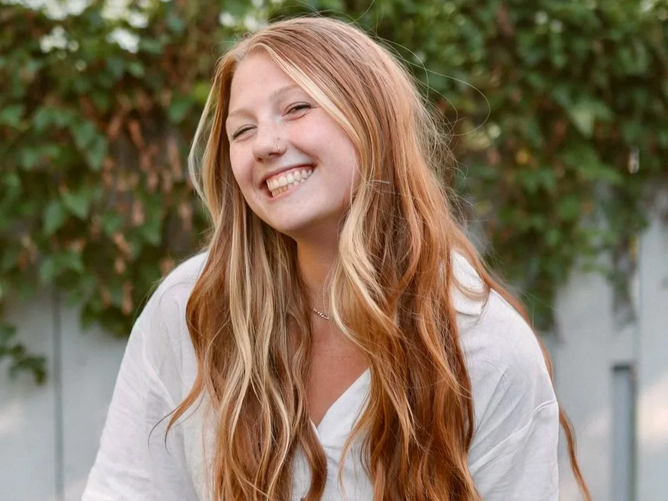 A woman with long, wavy red hair smiling outdoors with greenery in the background.