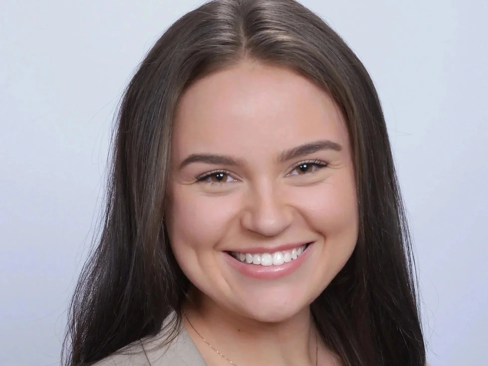 Close-up of a smiling young woman with long brown hair and brown eyes, showing her teeth, against a plain light background.