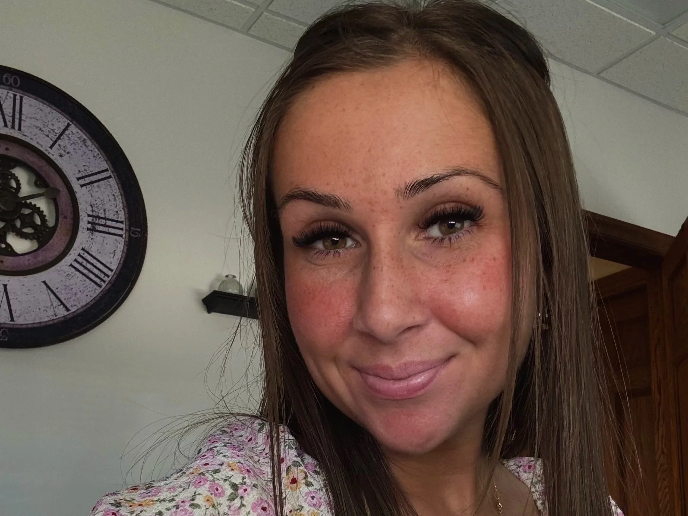 Close-up of a smiling woman's face showing her nose, lips, and eyes, with a wall clock partially visible in the background.