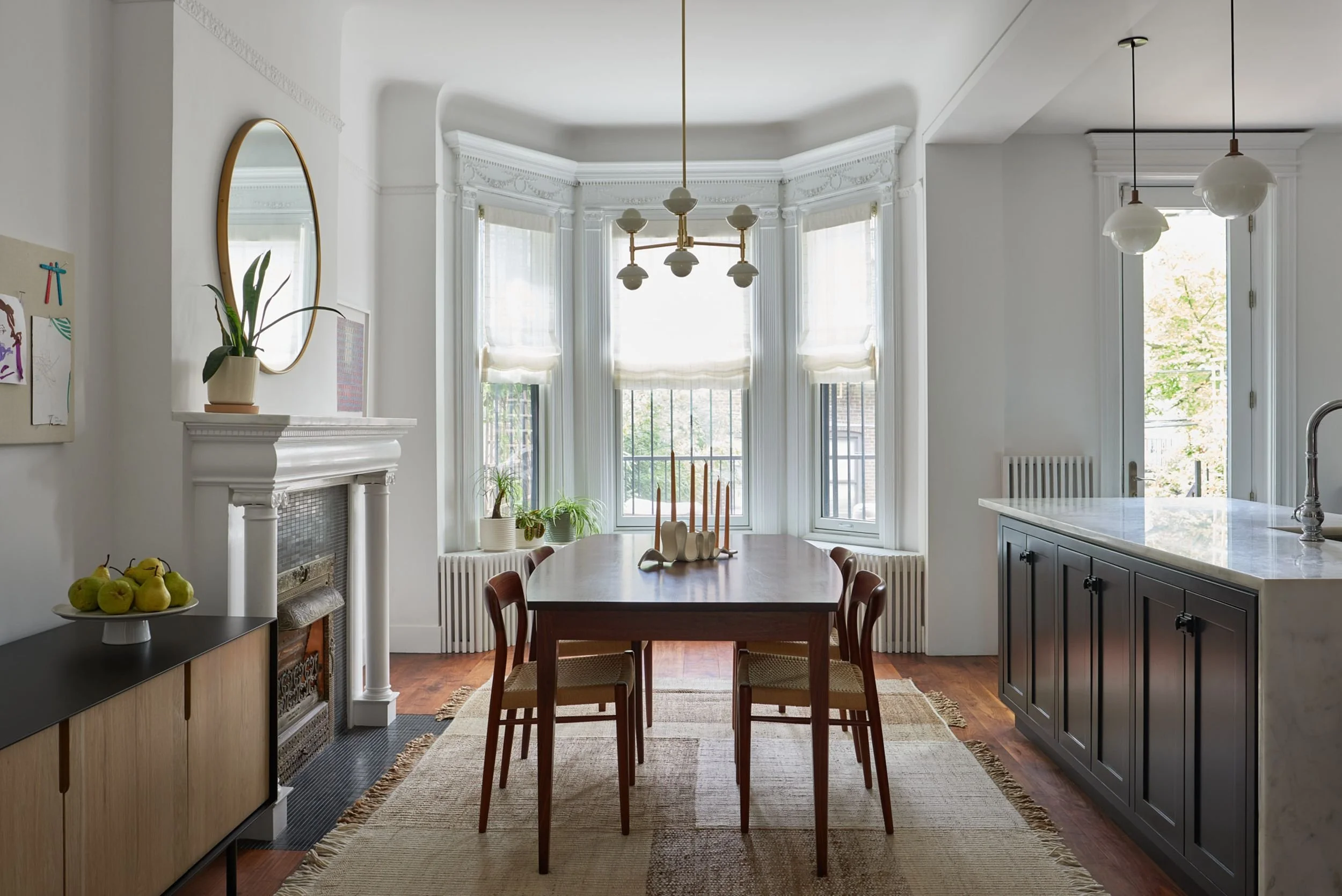Bright dining area with large bay windows, long oval wooden table, and six chairs. White walls and ceiling with decorative crown molding, a modern chandelier, and eps. Kitchen with dark cabinetry and marble countertop visible to the right. Plants and