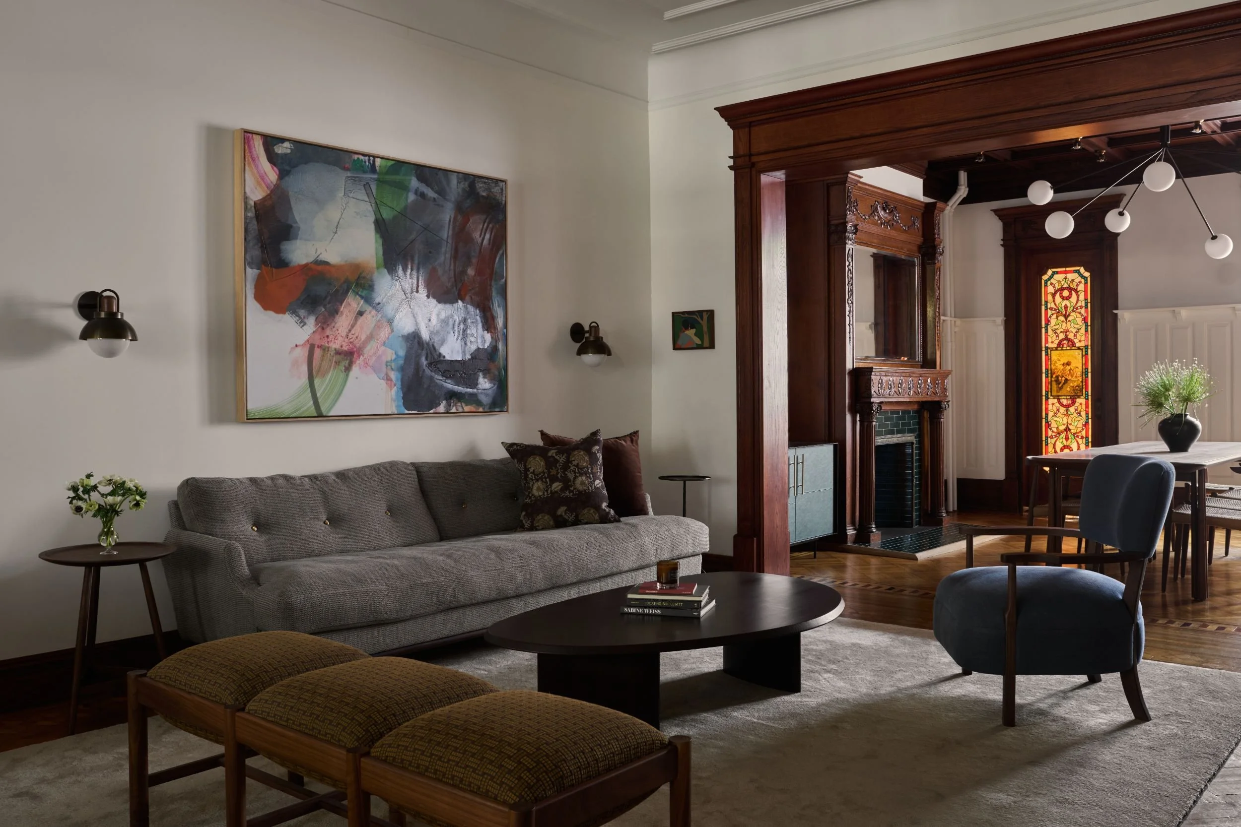A living room with a gray sofa, a small side table with a vase of flowers, framed abstract art on the wall, and wooden paneling enclosing a fireplace. There is a stained glass window in the background and a modern chandelier hanging from the ceiling.