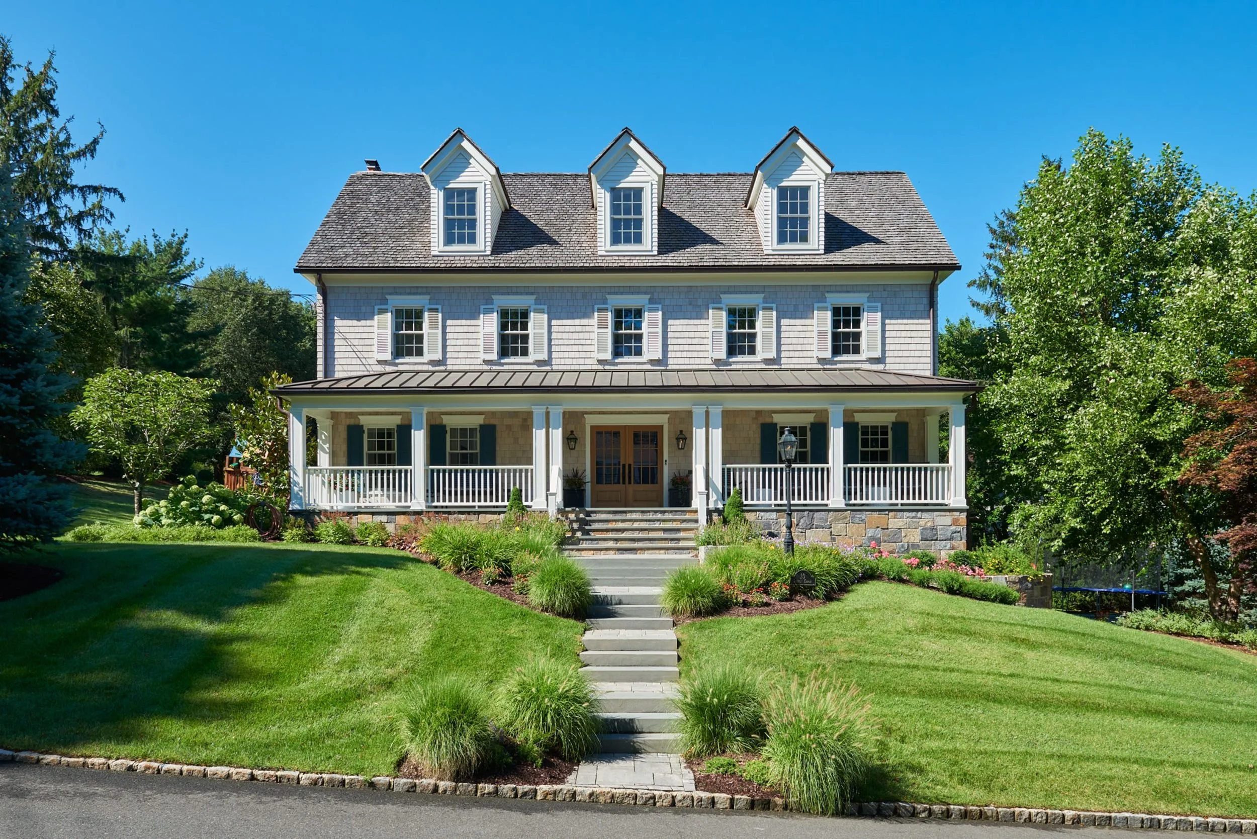 Front view of a large house with a porch, surrounded by a well-maintained garden with steps leading to the entrance, lush green trees, and a clear blue sky.