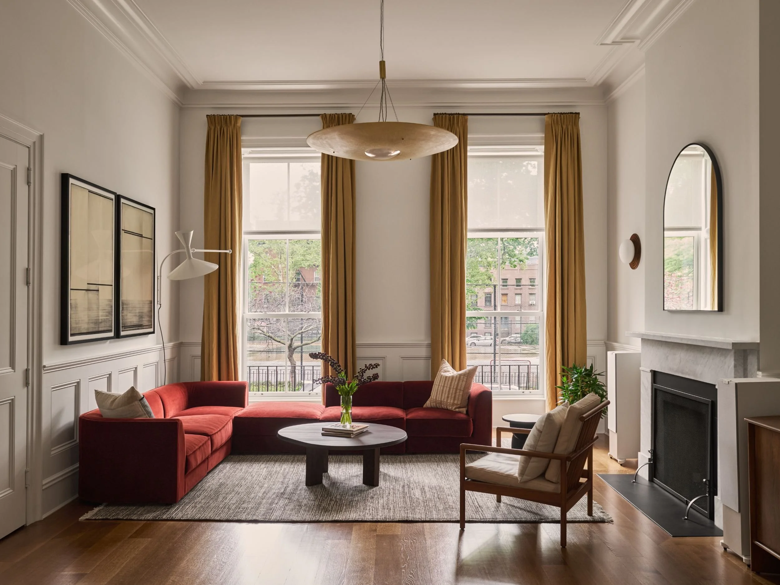 Living room with high windows, mustard-colored curtains, a red sectional sofa, a round coffee table with a vase of flowers, a beige armchair with throw pillows, and a fireplace with a mirror above.