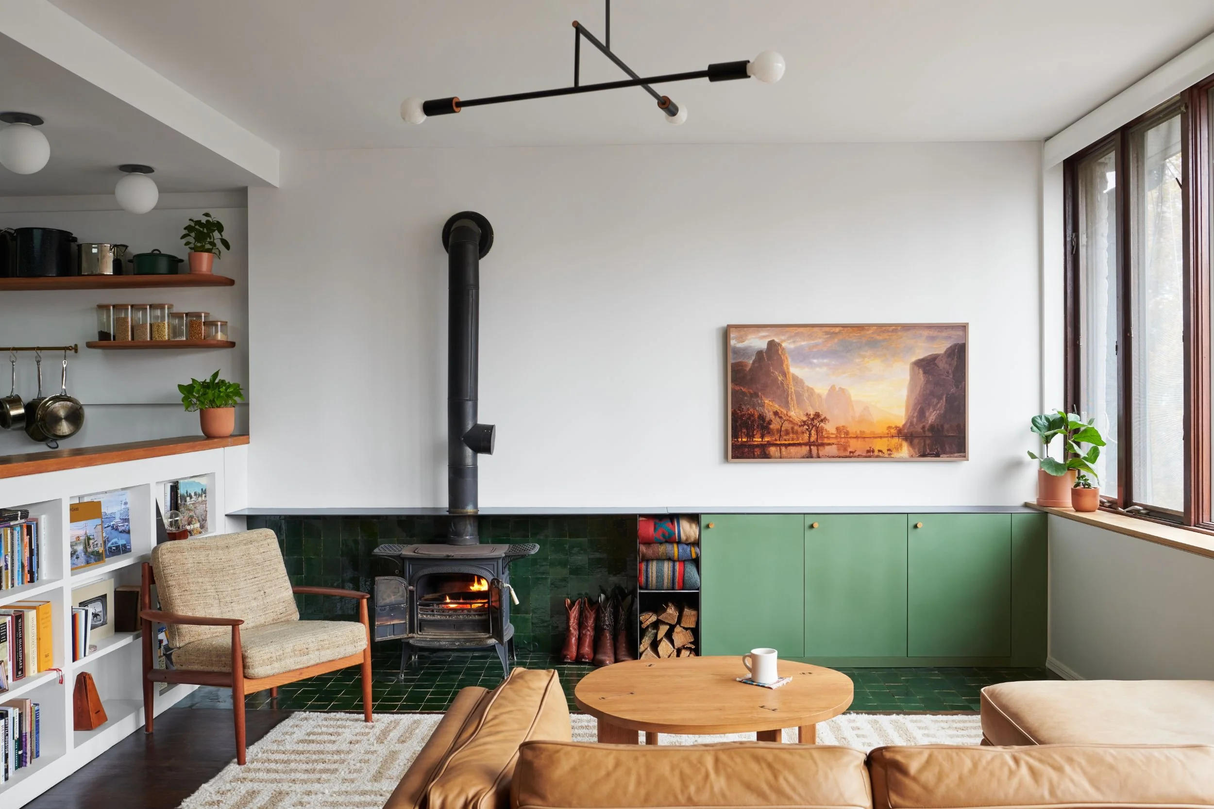 A cozy living room with a black stove, green tiled hearth, wooden shelves with jars, plants, a fireplace, framed landscape artwork, and large windows with sheer curtains.