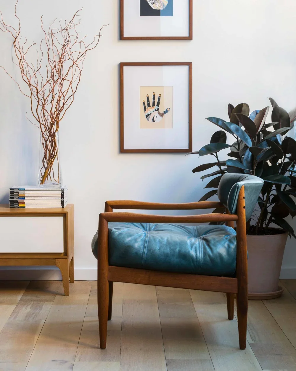 Interior living space with a wooden armchair with teal leather cushion, large potted plant, minimalist side table with stacked books, modern framed art on white wall, decorative dried branches in a glass vase.