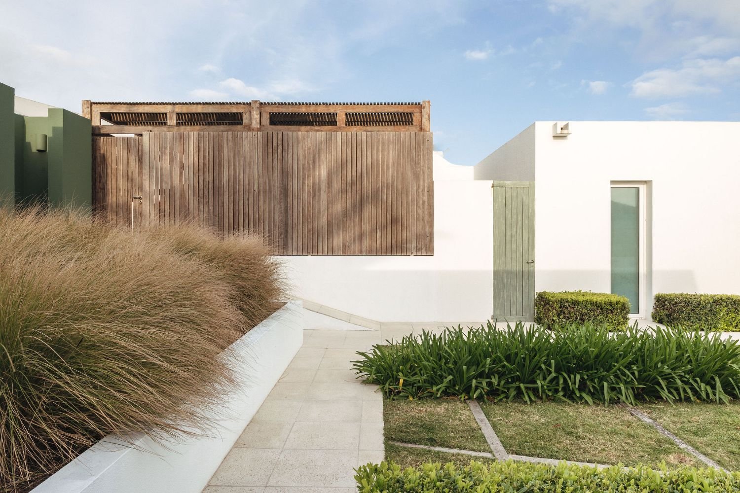 Modern house with white walls, a green garden door, and landscaped greenery, including tall grasses and trimmed bushes, under a blue sky.