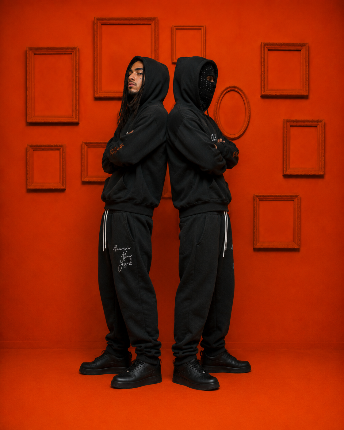 Two models wearing black MXNY Rebels Never Die hoodies and sweatpants, standing back-to-back in front of an orange wall for the Mavrix NY campaign.